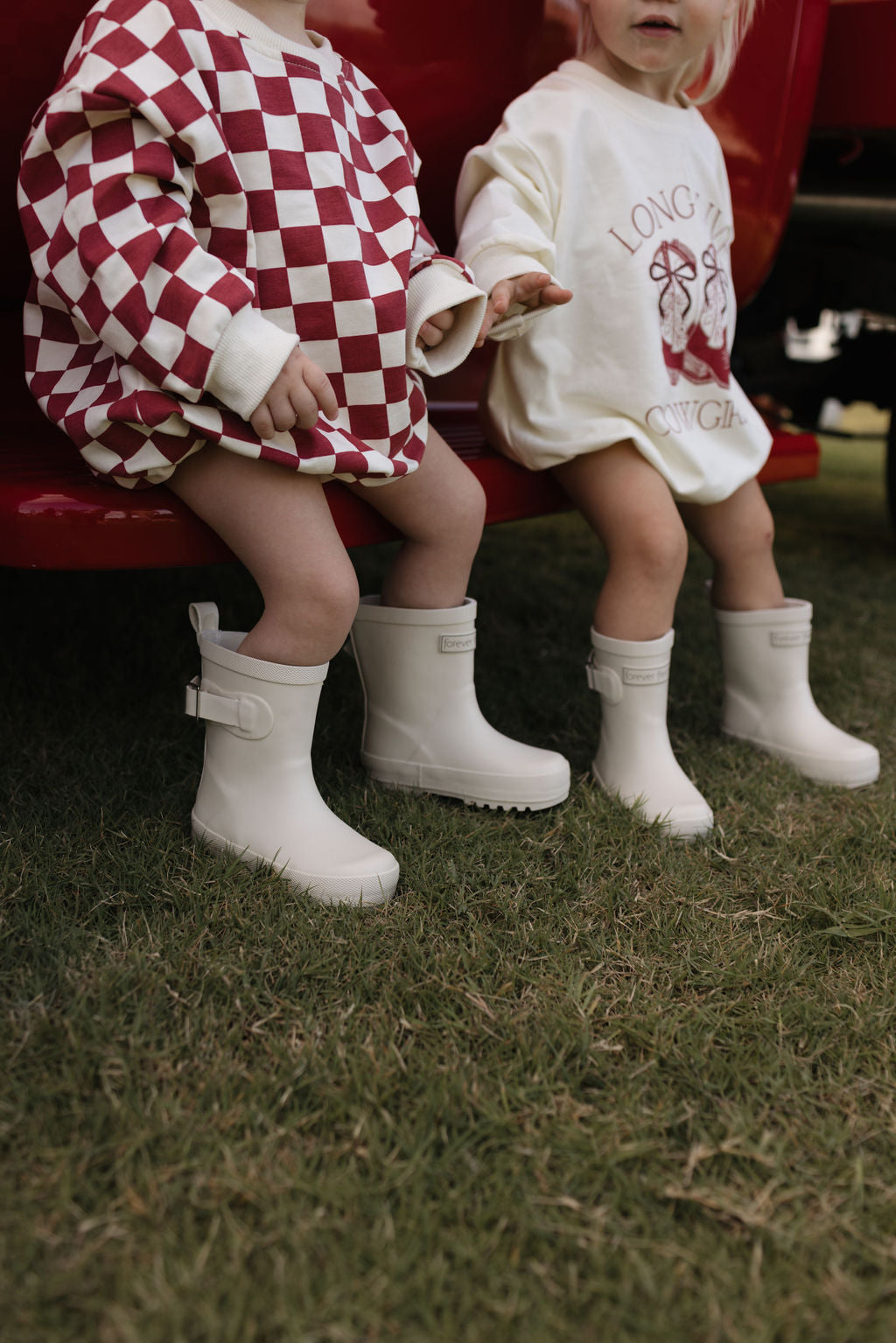 Two children sitting on a red surface, wearing white rain boots. One is dressed in a red and white checkered outfit, while the other is sporting the "Long Sleeve Romper | Long Live Cowgirls" by forever french baby, a cream sweatshirt romper featuring red text that’s perfect for the cooler months. They are outdoors on the grass, embracing the charm of carefree days.