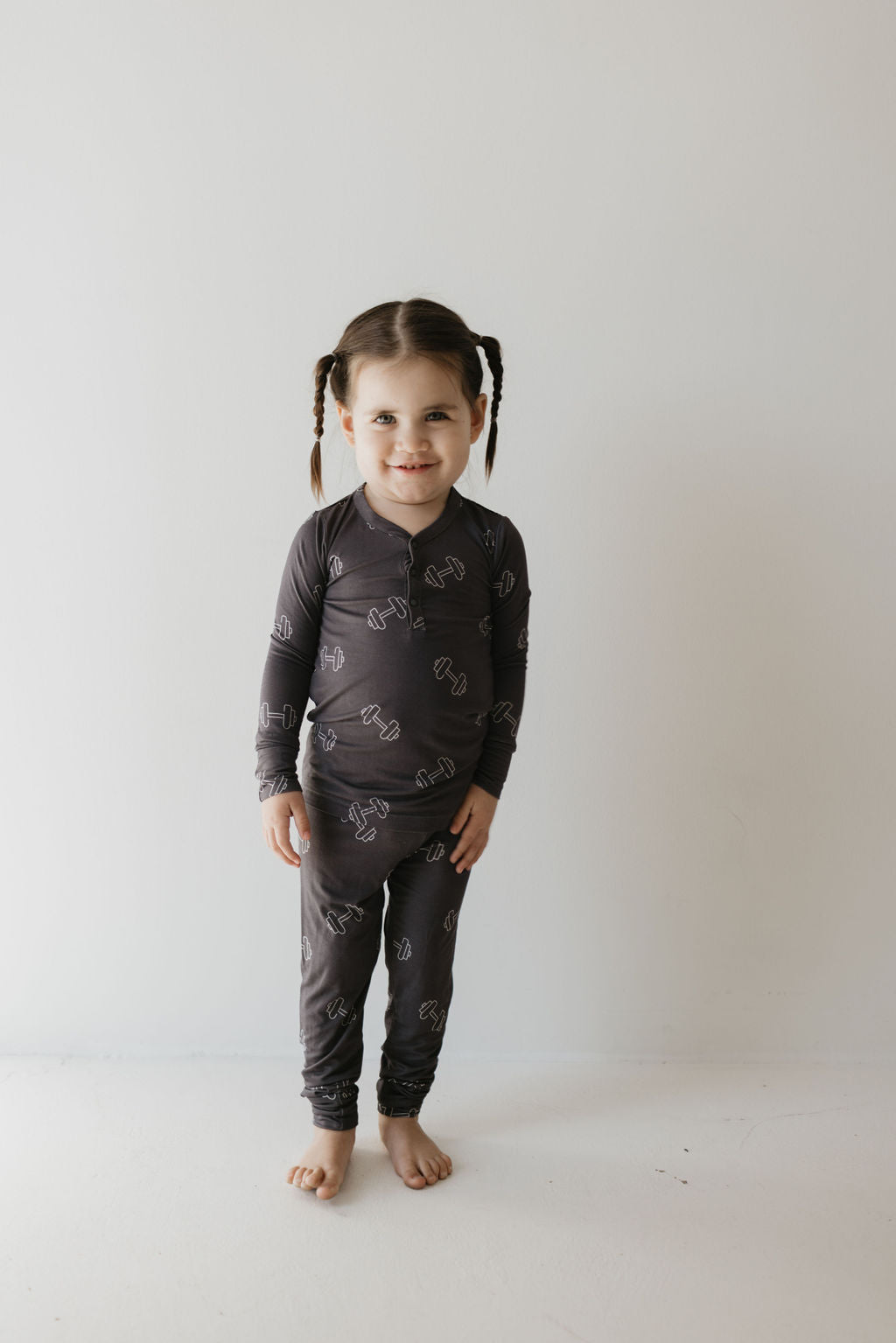 A young child with braided hair stands barefoot on a light floor against a plain wall, wearing matching dark "Arm Day" bamboo pajamas by forever french baby, featuring a geometric pattern. Their gentle smile aimed at the camera is complemented by the breathable sleepwear.