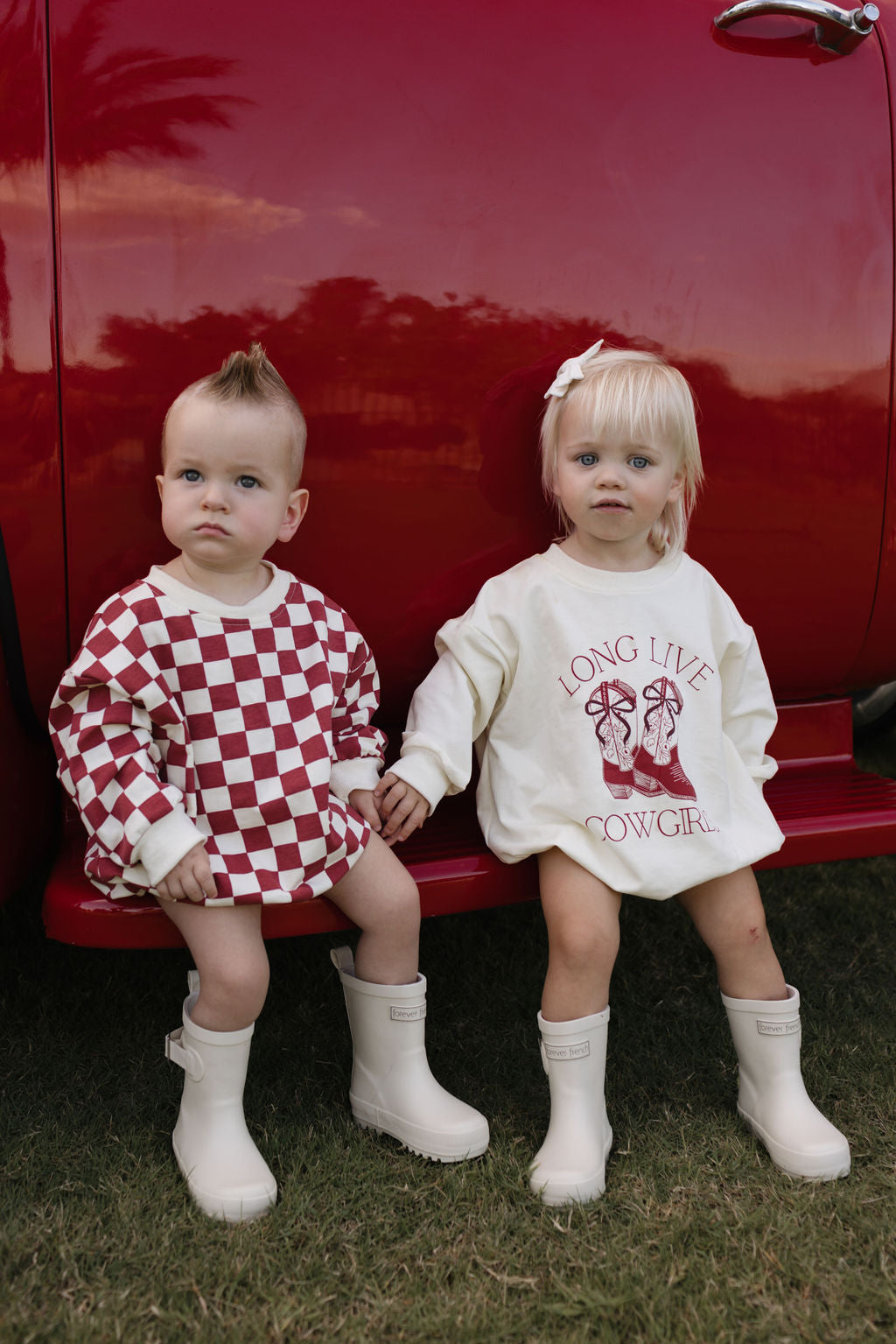 Two toddlers sit on the step of a red vehicle, dressed for the cooler months. One wears a red and white checkered outfit, while the other sports The Quinn Long Sleeve Romper from lolo webb, featuring "Long Live Cowgirl" printed on it. Both have light hair, are wearing white boots, and fall within the newborn to 24-month age range. Grass peeks beneath them.