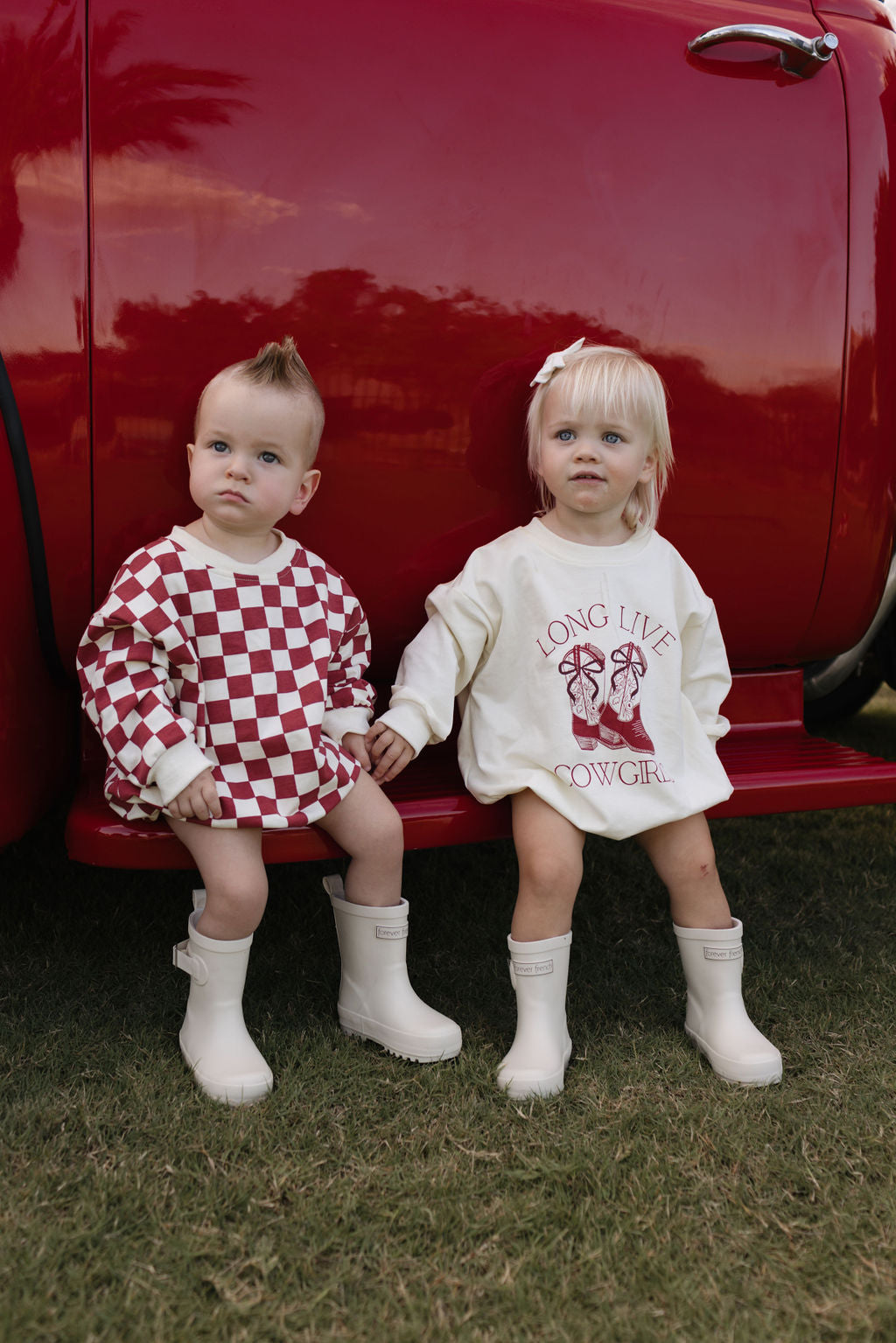 Two toddlers are seated on the back of a red truck. One is wearing a checkerboard-patterned outfit with boots, while the other is dressed in the "Long Sleeve Romper | Long Live Cowgirls" by forever french baby, paired with matching boots. Both have light hair and are enjoying the cooler months as they relax on the grass.