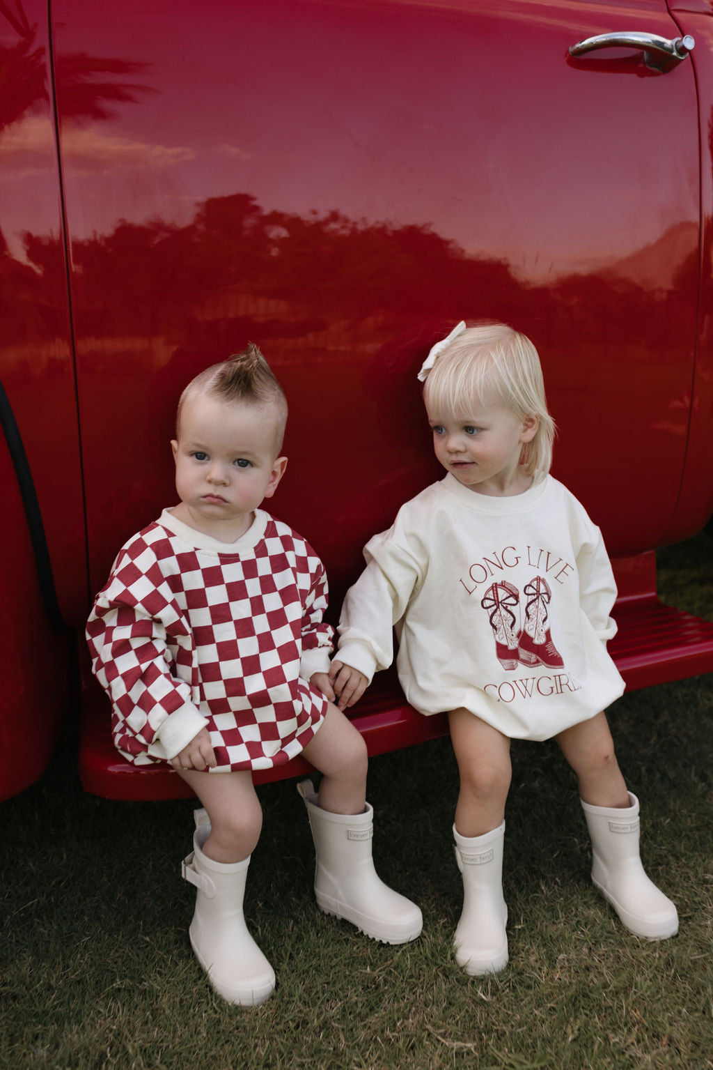 Two toddlers sit beside a red vehicle. One is dressed in a red and white checkered outfit, while the other wears the cozy "**Long Sleeve Romper | Long Live Cowgirls**" from **forever french baby**, ideal for cooler months. Both toddlers have short, light-colored hair and are sporting white boots, enjoying their time on the grass.