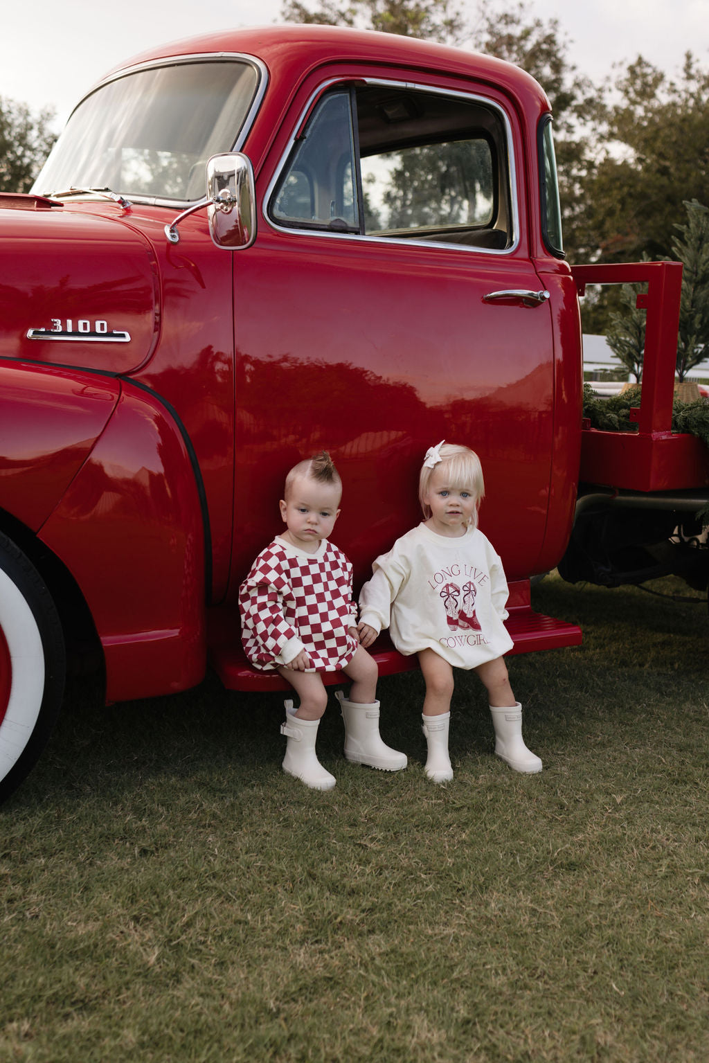 Two toddlers, dressed for the cooler months, sit on the step of a vintage red truck. One wears a red and white checkered outfit, while the other sports a cozy Long Sleeve Romper called "the Quinn" from lolo webb. Both have white boots on their tiny feet. Behind them stands a tree with grass underfoot.