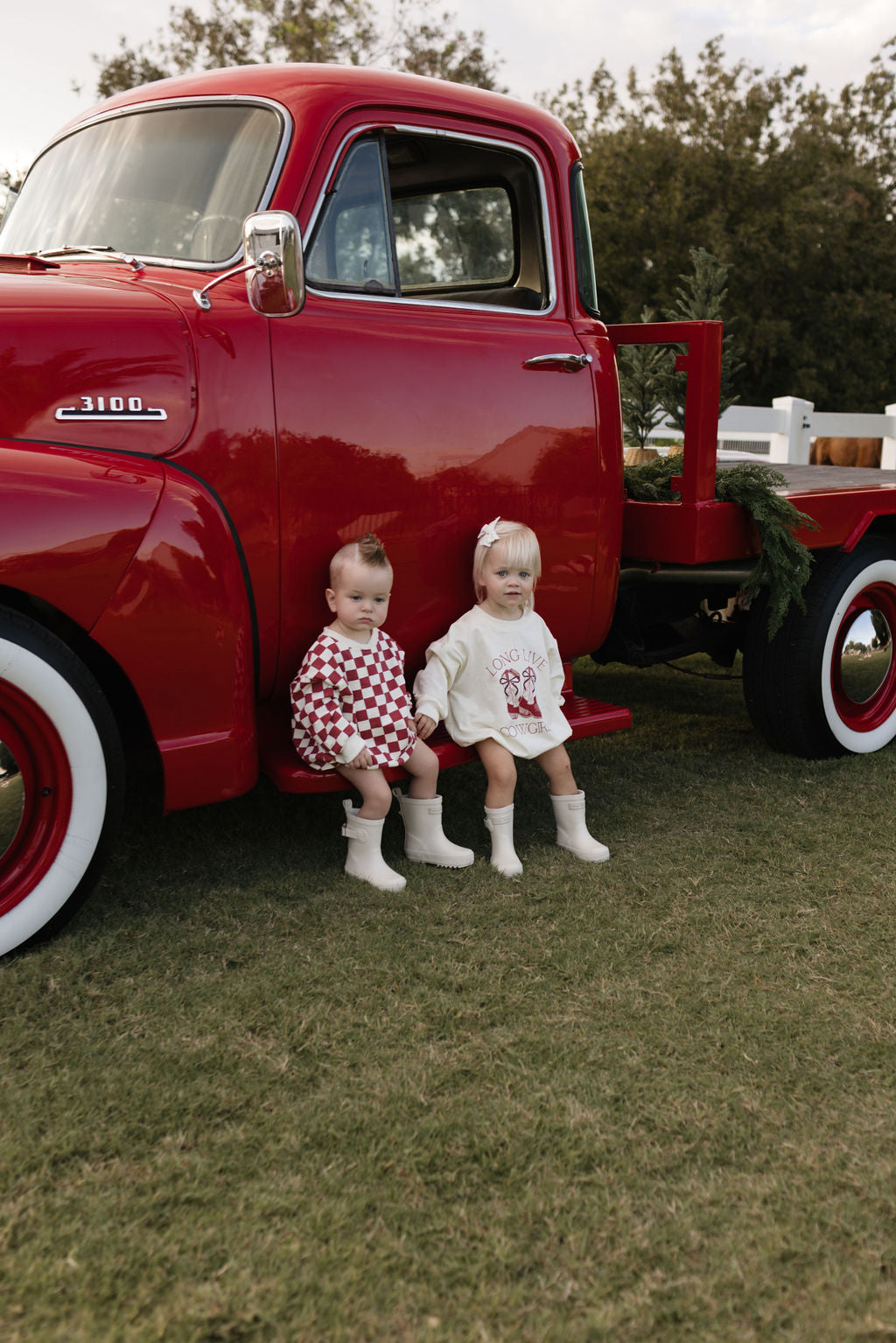 Two young children in festive outfits sit on the step of a vintage red truck parked on a grassy field, with a fence and greenery enhancing the nostalgic atmosphere. In the cooler months, a Long Sleeve Romper from the Quinn collection by lolo webb would be ideal for little ones enjoying such cozy scenes.