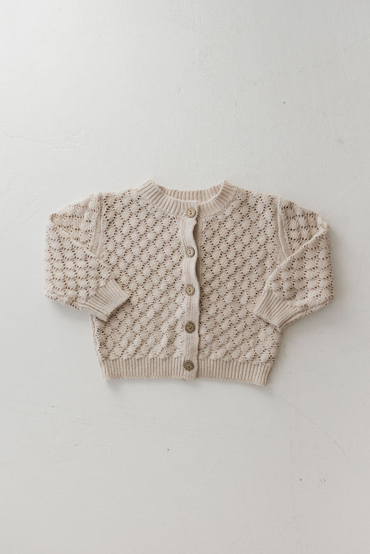 A cozy Knit Cardigan | Biscuit by forever french baby is displayed flat, showcasing its warm-toned cream hue and delicate diamond pattern. Made from breathable cotton, it has small buttons along the front and ribbed details on the cuffs and hem. The background is a plain, light-colored surface.