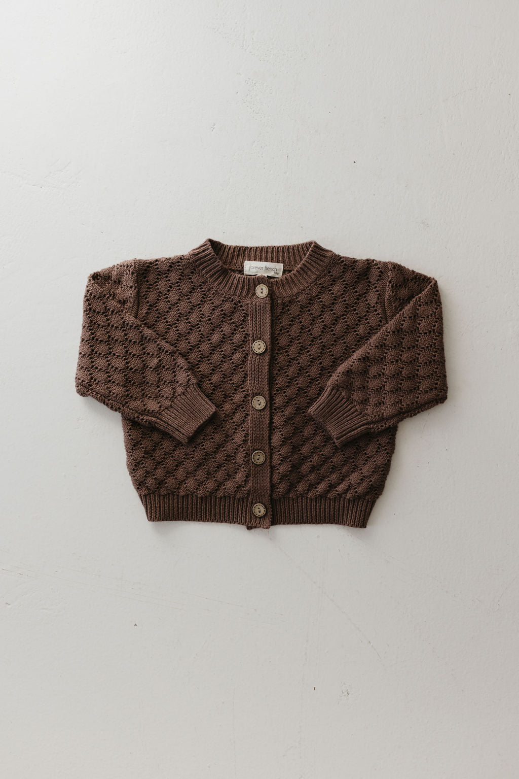 Displayed on a light background is the Knit Cardigan | Clove, from forever french baby, in a brown hue. This knitted cardigan is crafted from breathable cotton and showcases a textured pattern with wooden buttons. It features long sleeves and a simple round neckline, making it perfect for versatile styling.