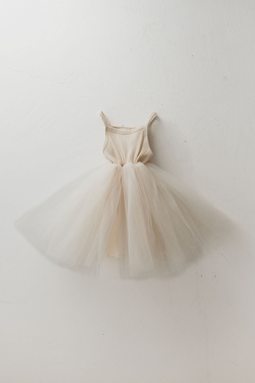 The ff Tutu Dress | Sugar by forever french baby is a cream-colored kids' wardrobe staple, showcasing a fitted ribbed bodice and a fluffy tulle skirt that hangs gracefully against a white wall.