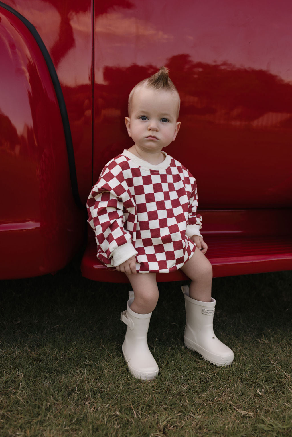 A baby with a mohawk hairstyle perches on the edge of a vibrant red vehicle, dressed in the cozy Long Sleeve Romper by lolo webb, featuring a charming red and white checkered pattern ideal for cooler months. Their white boots rest on the grass as they gaze directly at the camera—a perfect look for newborns to 24 months.