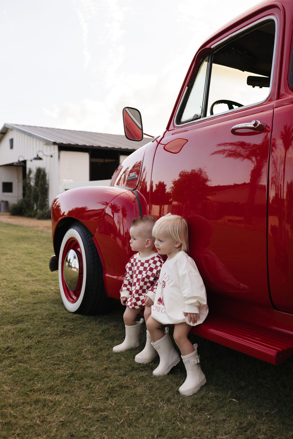 Two children in matching outfits and boots sit on the side step of a vintage red truck, dressed warmly for the cooler months. A barn stands in the background with grassy fields beneath them, while partly cloudy skies hover above. One wears a cozy Long Sleeve Romper from lolo webb's "the Quinn" collection, perfect for newborns to 24 months.