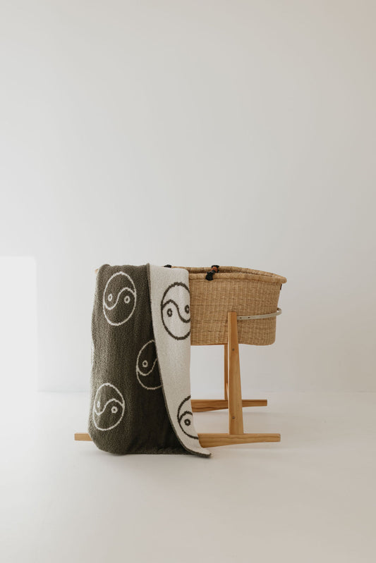 A woven baby bassinet from forever french baby rests on a wooden stand in a minimalist, light-filled room. A Plush Blanket | Sage Yin Yang with black and white yin-yang symbols drapes over the side. The background is a soft cream color, creating a serene atmosphere.