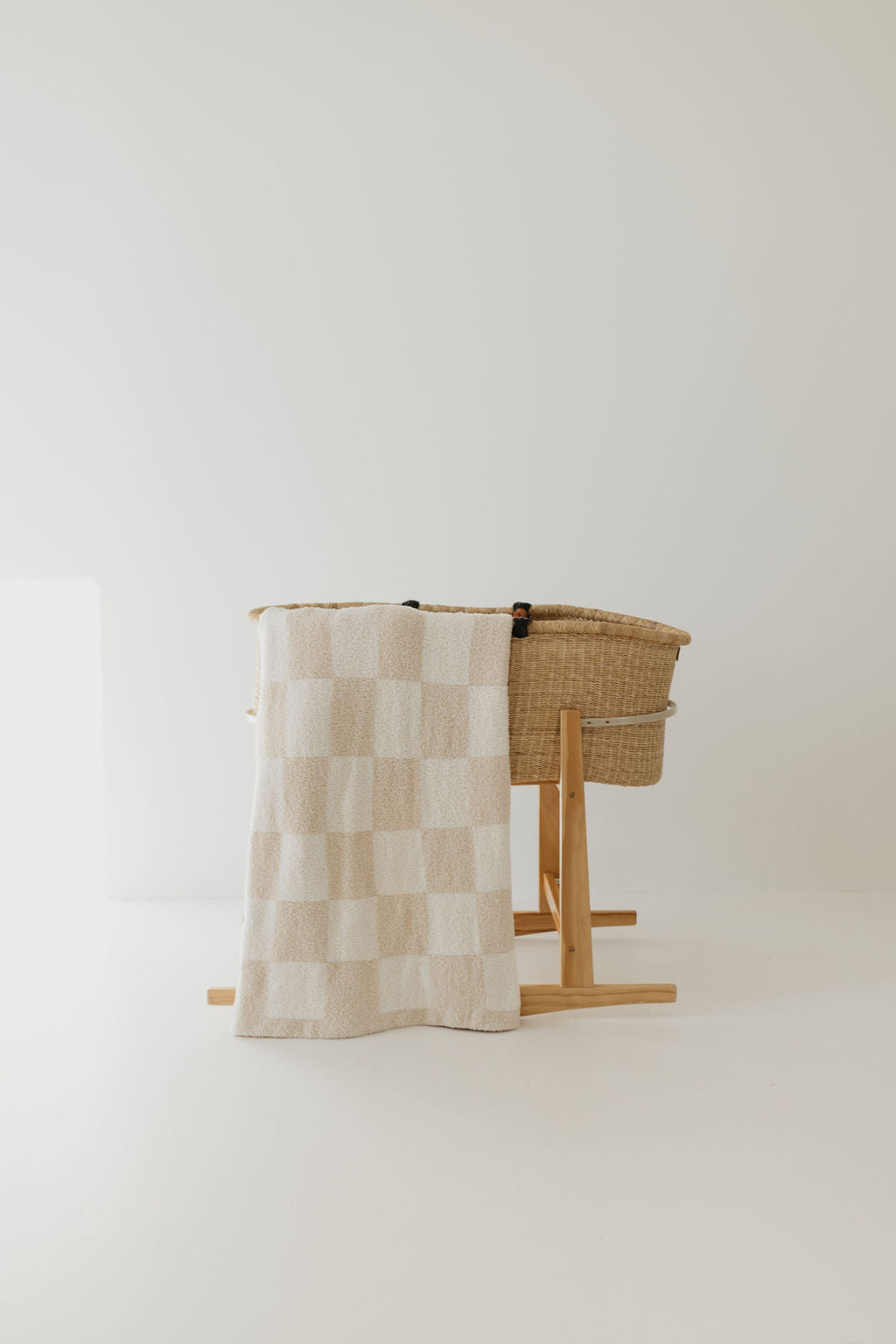 A wicker bassinet with a wooden stand is set against a light background. A dreamy, soft Plush Blanket in the Cream Checkerboard pattern from forever french baby is draped over the side. The scene remains minimal and serene, conveying a sense of calm and simplicity.