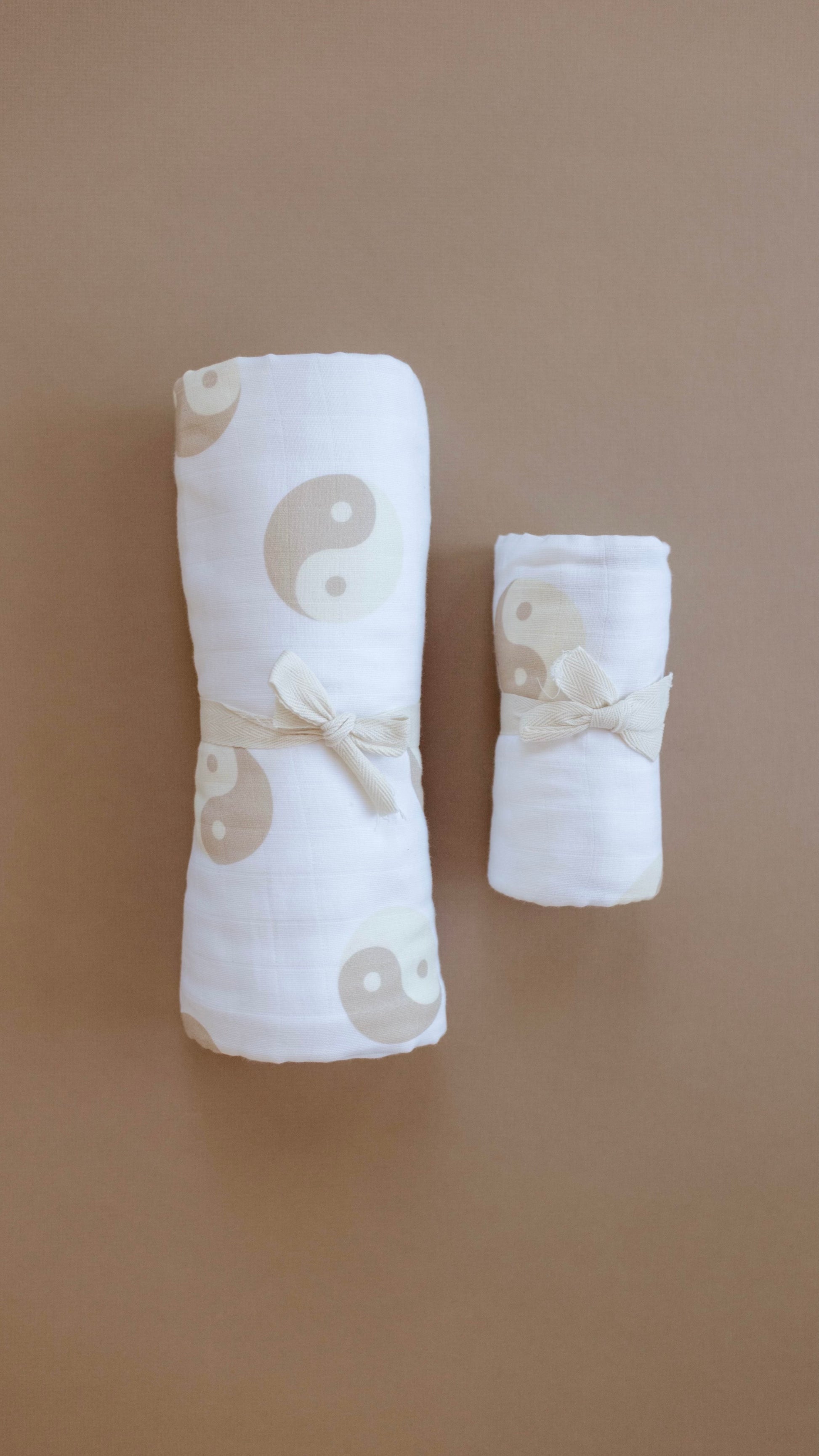 Two pieces of the "Muslin Swaddle | Tan & White Yin Yang" by forever french baby, each rolled up and tied with a beige bow, are made from muslin cotton. The fabric showcases a tan and white yin-yang pattern. The swaddles are placed on a plain brown background, with one roll larger than the other.