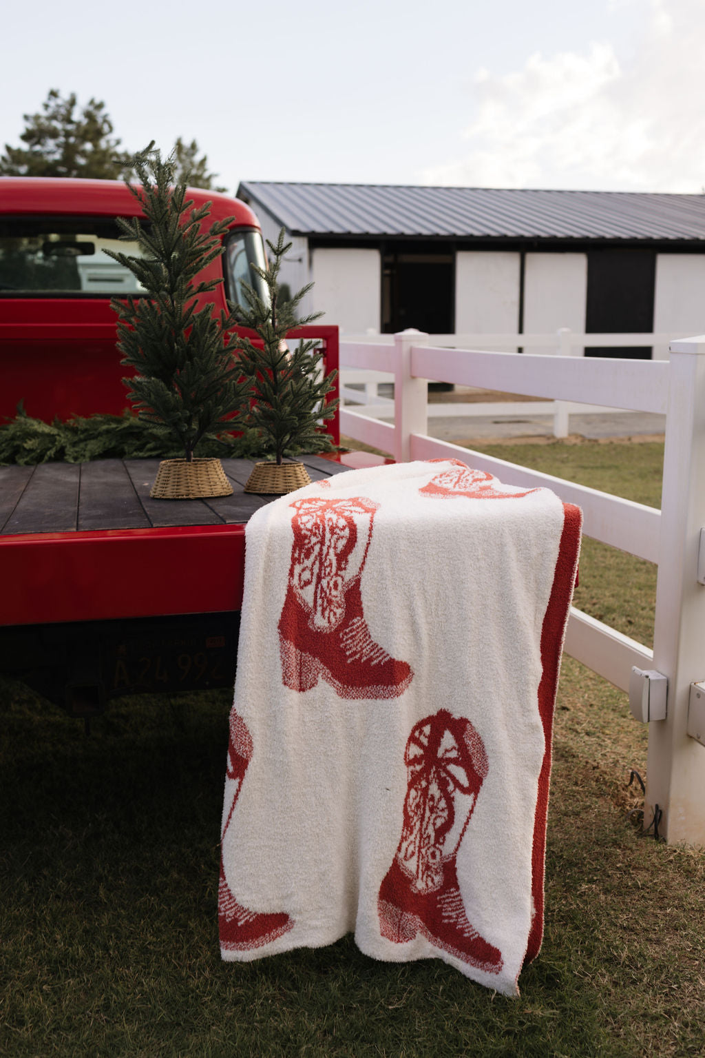 A red truck parked by a white fence holds a small potted evergreen tree. Draped over the tailgate is the lolo webb Plush Blanket, featuring dreamy red cowboy boot designs from their Cowgirl Christmas collection. In the background, a barn with a metal roof completes this picturesque scene.