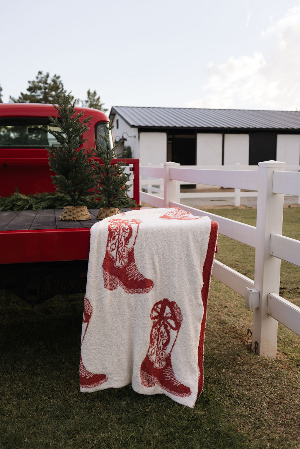 A red truck parked near a white fence with a small evergreen tree in the back proudly displays the dreamy Plush Blanket | Cowgirl Christmas by lolo webb, featuring red cowboy boots draped over it. In the background, a white building with a dark roof and open windows completes this serene scene.