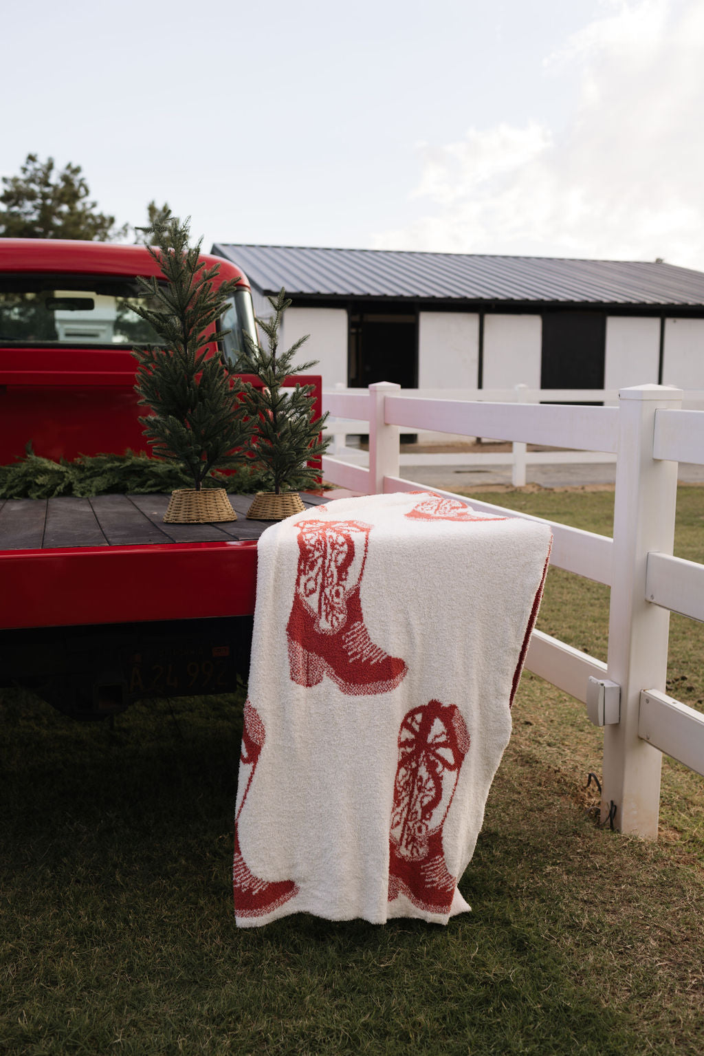 A red pickup truck with a small Christmas tree in the back is parked next to a white fence. A dreamy soft blanket from the lolo webb collection, adorned with festive red boot designs, is draped over the edge of the truck. In the background, a barn completes this serene winter scene.