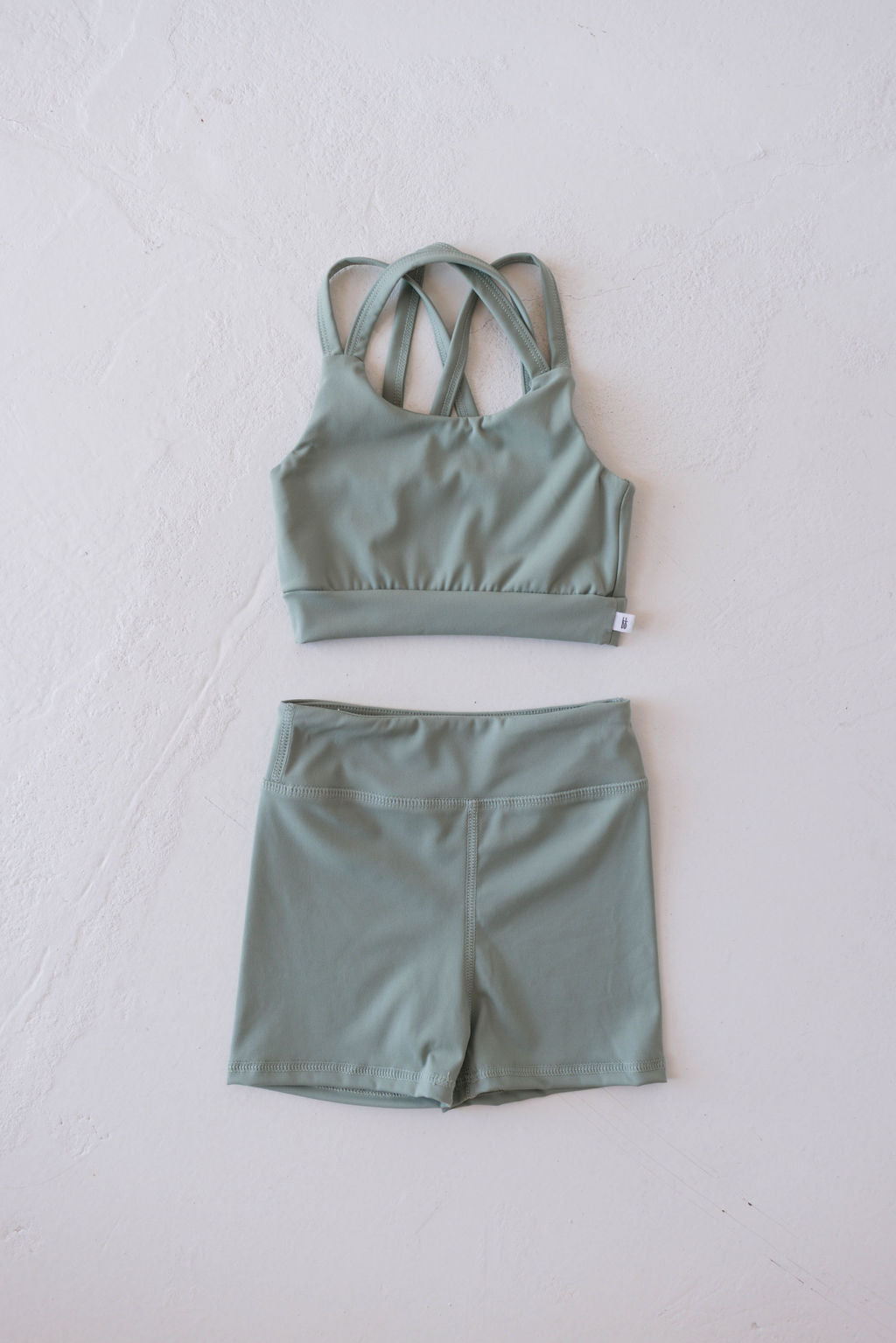 A light green children's workout set from forever french baby, featuring a sports bra with multiple crisscross straps on the back and a pair of high-waisted shorts, is laid out flat on a white textured surface.