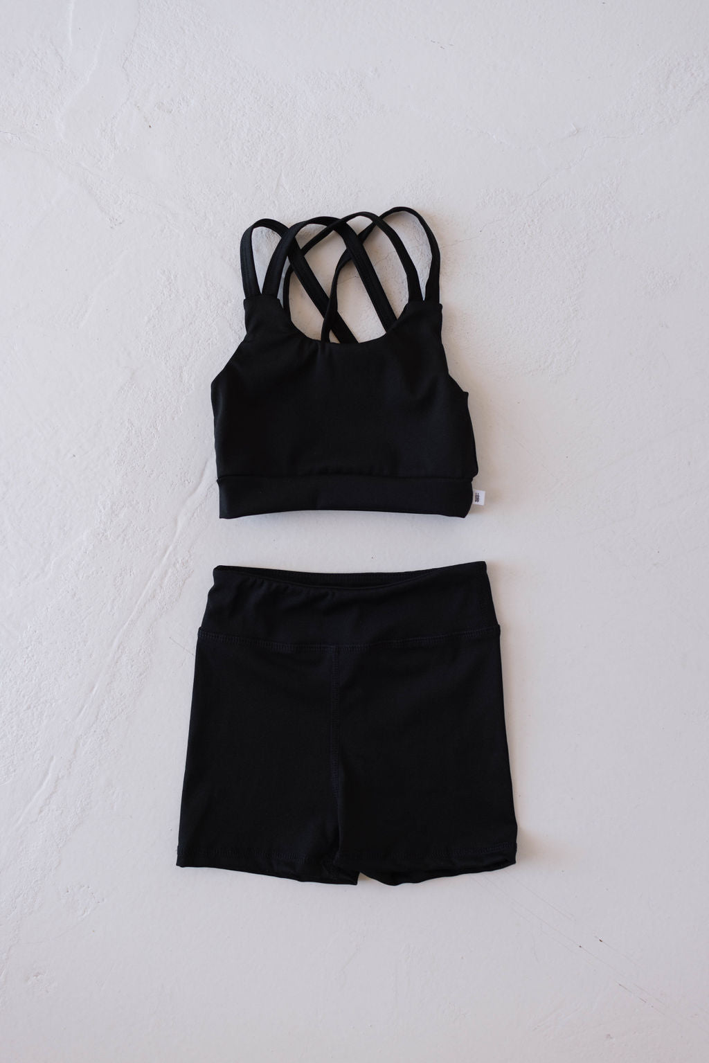 The forever French baby Children's Workout Set in black, showcasing a sports bra with multiple criss-cross straps and matching athletic shorts, is neatly laid out on a light-colored textured surface.