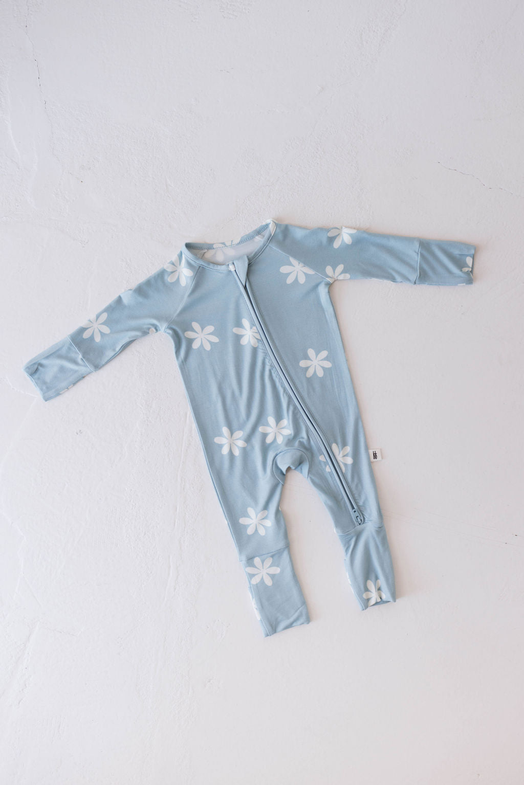 A light blue Bamboo Zip Pajamas by forever french baby, featuring the Daisy Blues pattern with white flowers. These eco-friendly pajamas have long sleeves, long legs, and a front zipper running from the neck to the left ankle. The onesie is laid flat against a white background.