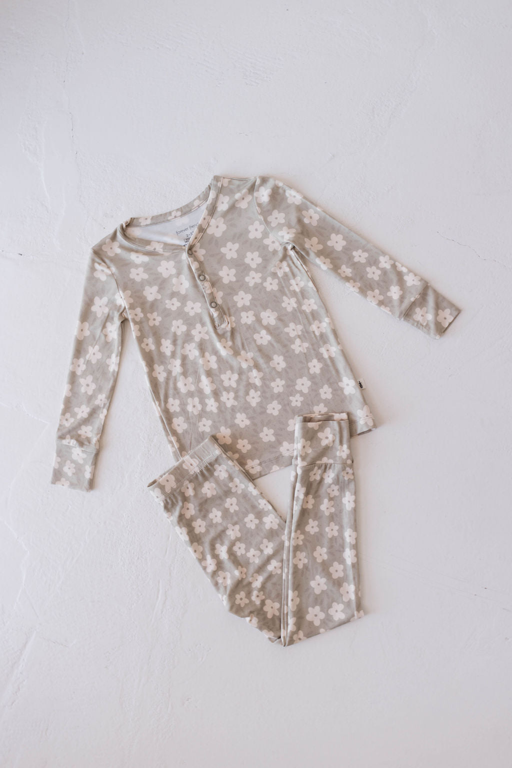 A matching two-piece pajama set from forever french baby lies flat on a white background. This Bamboo Two Piece Pajamas | French Gray Floral set, made from premium bamboo fabric, features an all-over pattern of white flowers. The top has long sleeves and three front buttons, while the bottoms have an elastic waistband.