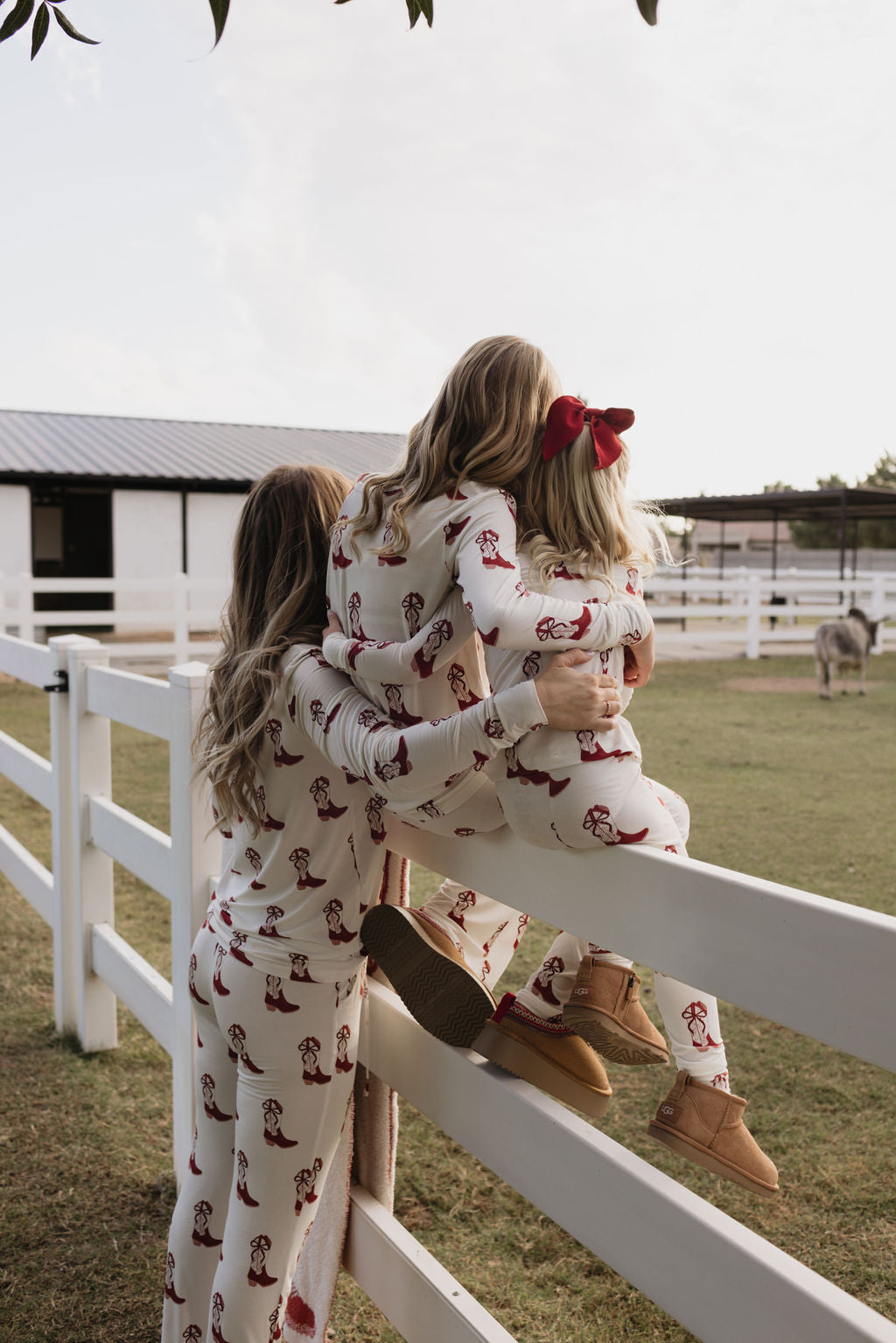 A woman and two children, all dressed in matching Bamboo Two Piece Pajamas from lolo webb's Cowgirl Christmas collection, stand against a white fence gazing at an outdoor scene. The child with a red bow delightfully looks towards a donkey in a paddock. Their breathable sleepwear enhances comfort as they take in the views of the barn and open sky.