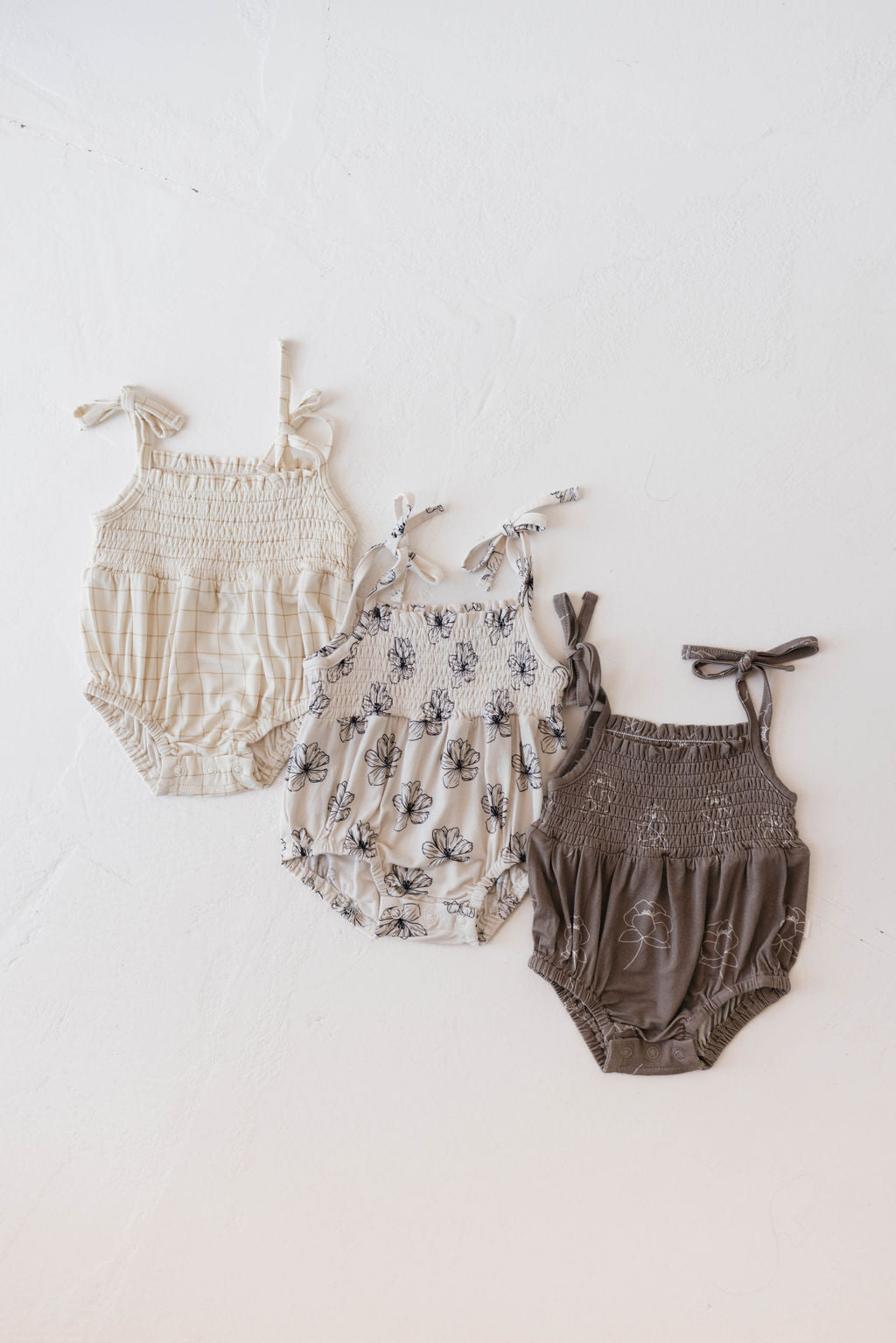 Three baby rompers laid out side by side. The first romper is a cream color with adjustable tie shoulder straps and a smocked bodice. The second is the Desert Bloom floral pattern from forever French baby, featuring thin straps. The third romper is a dark brown Bamboo Tie Top Romper from forever French baby, also with a smocked bodice and thin straps.