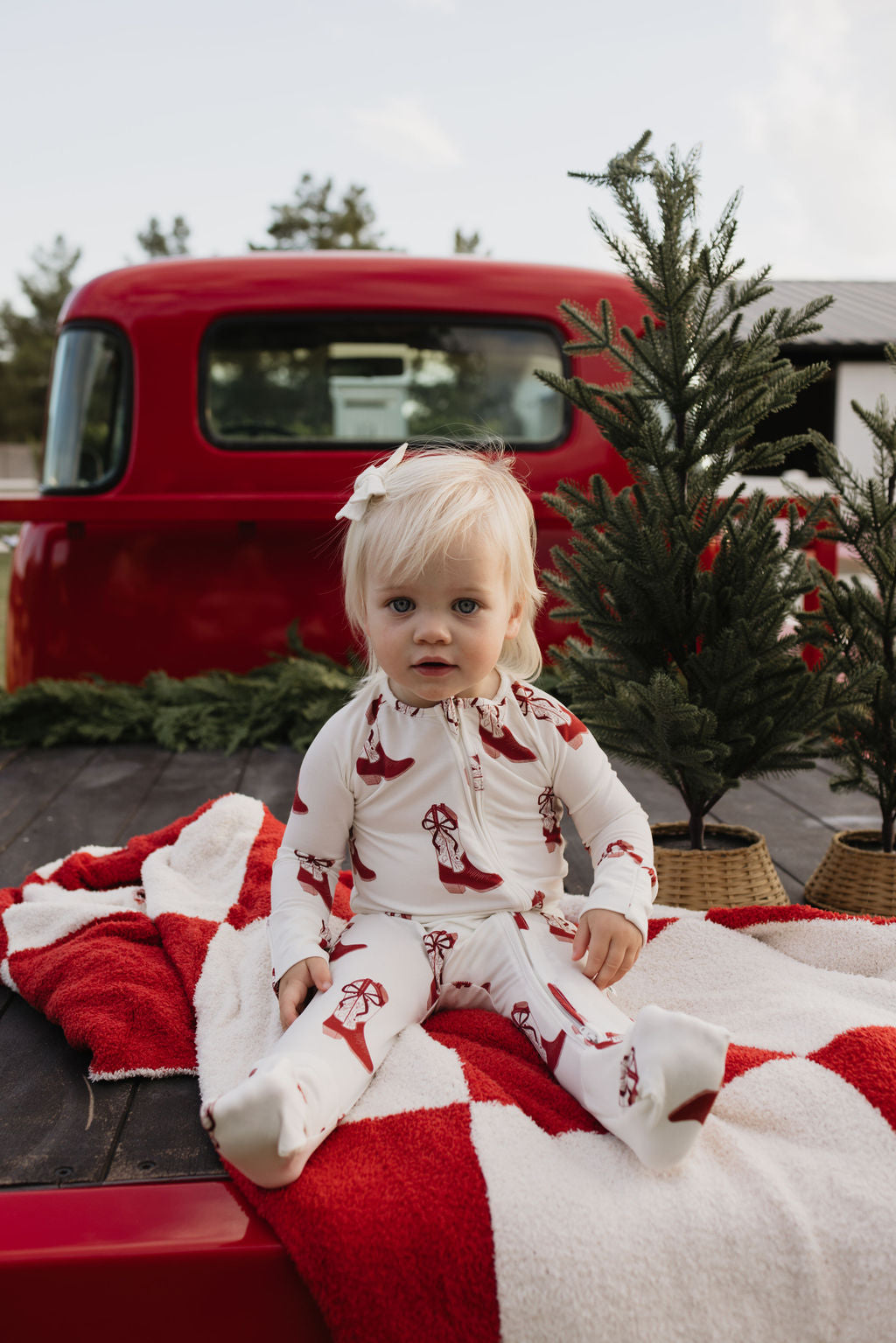 A toddler with blonde hair sits on a red and white blanket in the back of a truck, wearing cozy Cowgirl Christmas bamboo pajamas by lolo webb. Behind them are small potted pine trees, and part of a red truck is visible.