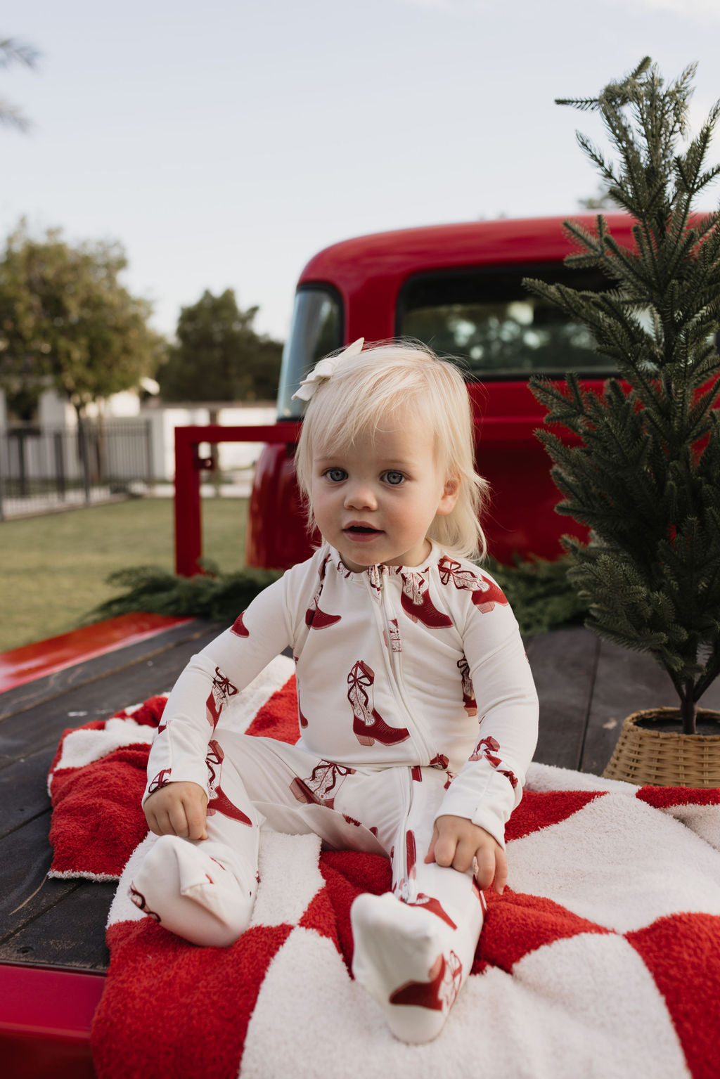 A toddler with blonde hair sits on a red and white blanket in the back of a red truck. The child is wearing lolo webb's Bamboo Zip Pajamas in Cowgirl Christmas, featuring breathable fabric and festive red patterns. A small evergreen tree in a basket is next to them, with trees and a fence visible in the background.