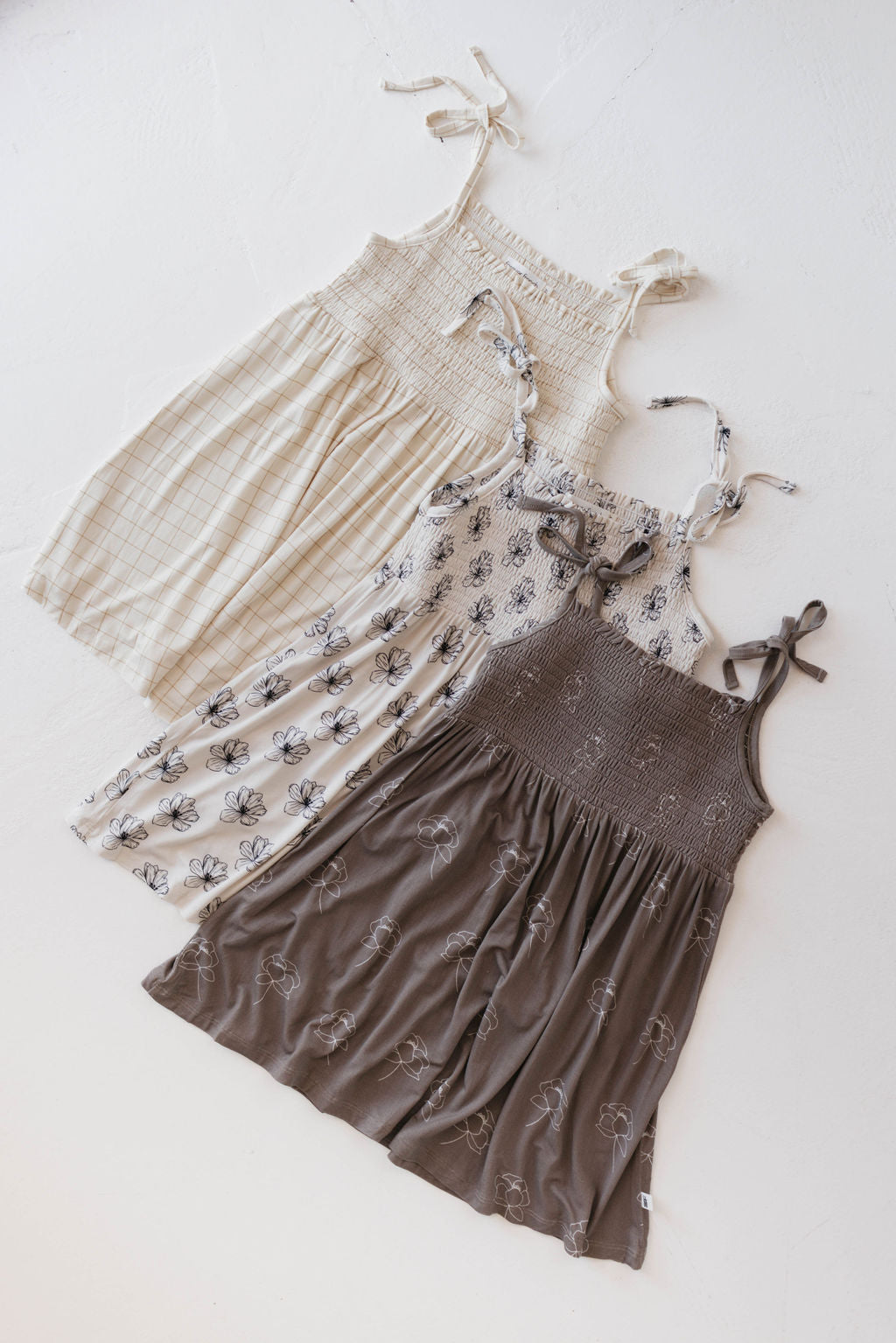 Three sleeveless smocked dresses with tie straps are laid out. The first is a comfortable dress in a light plaid pattern, the second is the Bamboo Tie Top Dress in Sweet Dreams Floral by forever french baby, and the third is dark brown with a matching floral design.