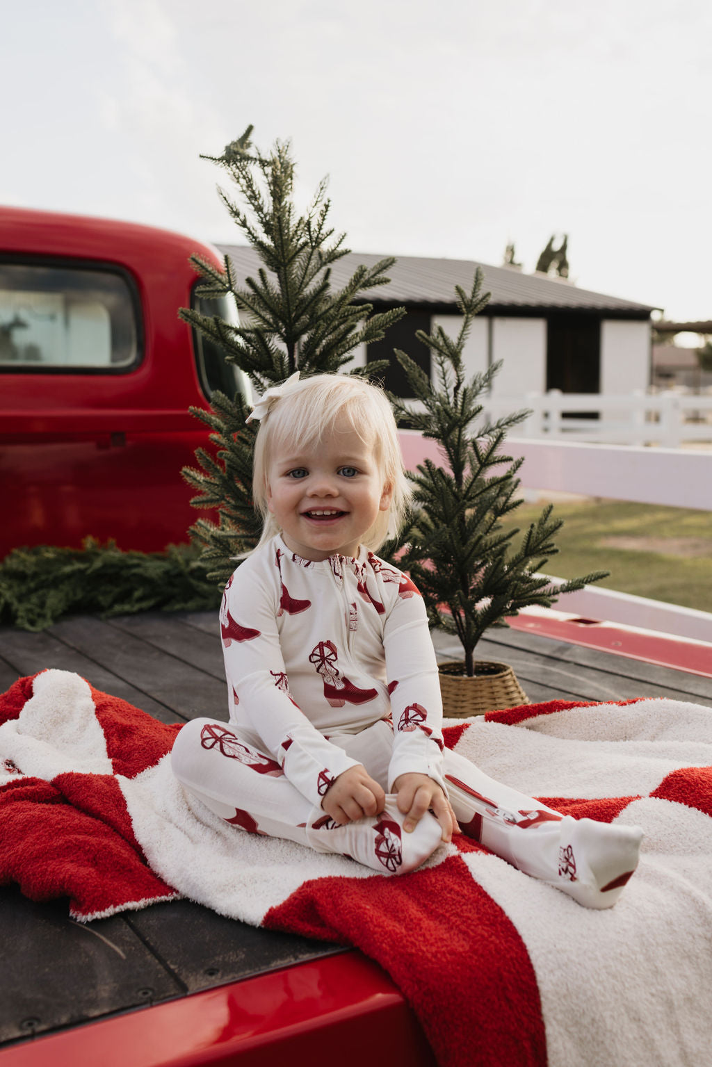 A smiling child with blonde hair sits on the back of a red truck, wearing 'Bamboo Zip Pajamas | Cowgirl Christmas' by lolo webb. The truck bed is adorned with small evergreen trees and a red and white blanket, creating a festive atmosphere.