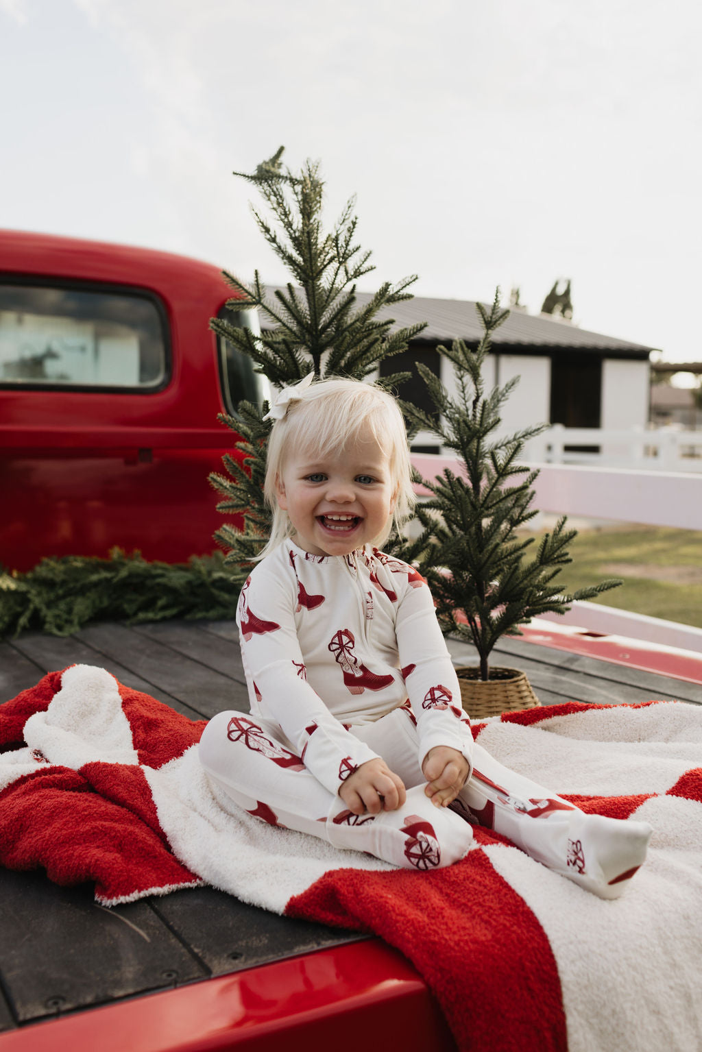 A smiling child with blonde hair sits on a red and white blanket in the back of a red truck, dressed in lolo webb's Bamboo Zip Pajamas featuring the Cowgirl Christmas design. Two small pine trees nearby enhance the holiday atmosphere.