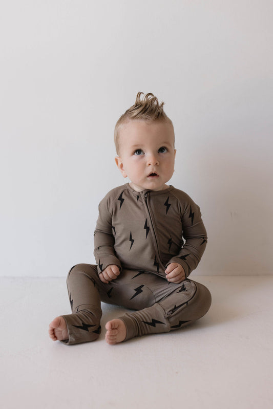 A baby in brown and black lightning bolt Bamboo Zip Pajamas from forever french baby sits on the floor against a plain white background, looking slightly upward. The baby, with light hair styled in a small mohawk, appears curious or attentive.