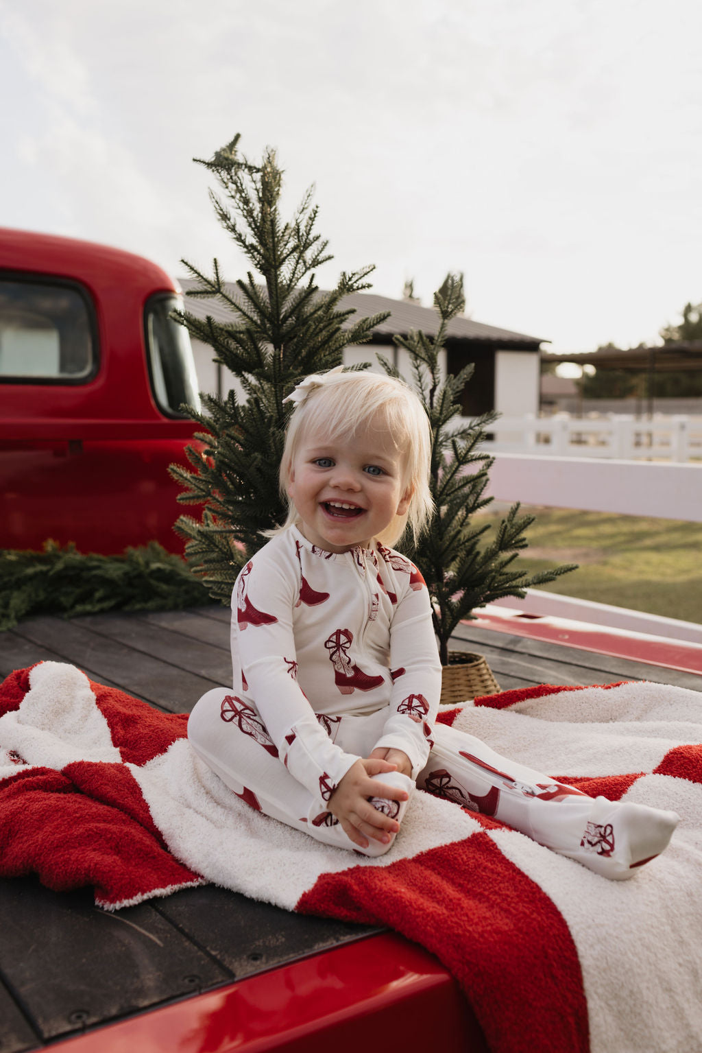 A cheerful toddler wearing lolo webb's Bamboo Zip Pajamas in the Cowgirl Christmas theme sits on the back of a red truck, surrounded by a small pine tree and a red-and-white blanket. The setting, featuring a barn and some fields, perfectly captures rural charm for cozy, breathable sleepwear moments.