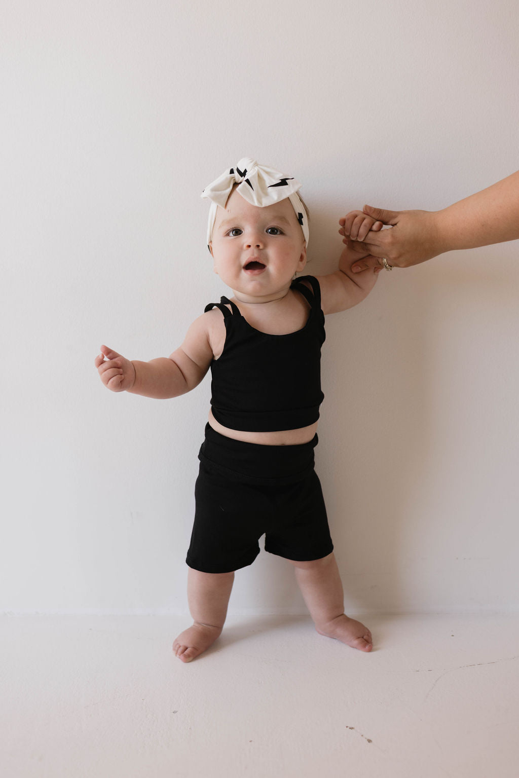 A baby, dressed in a black outfit and a Bamboo Head Wrap in the White + Black Bolt pattern from forever french baby, stands against a white background. An adult hand is gently holding one of the baby's hands for support.