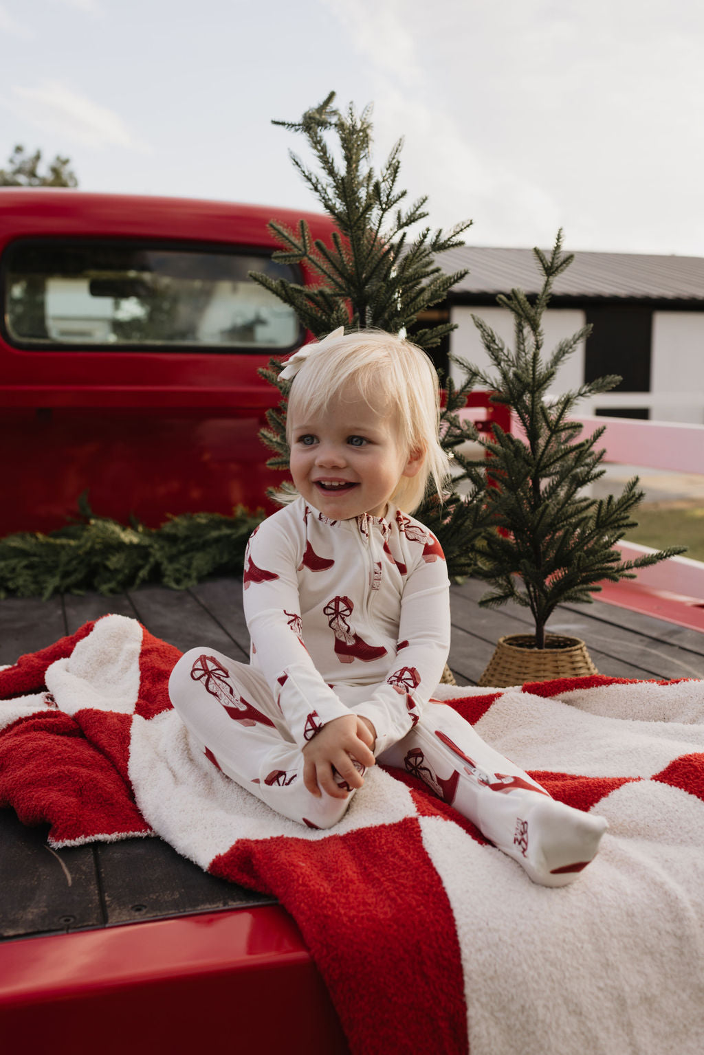 A joyful toddler, dressed in lolo webb's Bamboo Zip Pajamas | Cowgirl Christmas, sits on a red and white blanket in the back of a red truck. Surrounding small pine trees enhance the cheerful holiday atmosphere.