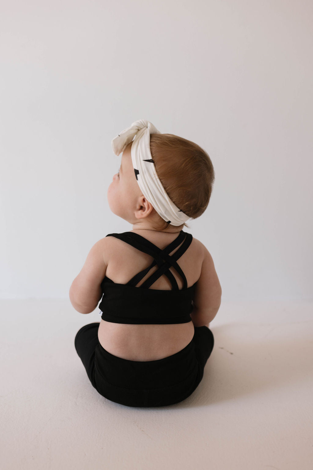 A baby sits on the floor, facing away from the camera, dressed in forever French baby's Children's Workout Set in black and a white headband with a bow. The stylish outfit showcases a crisscross back design against a plain, light-colored wall background.