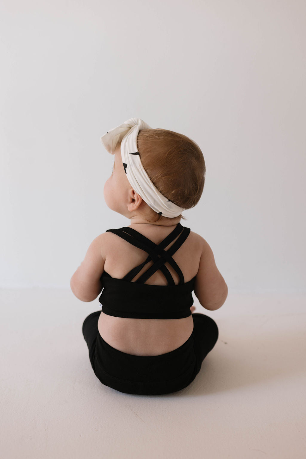 A baby is sitting and facing away from the camera, dressed in a forever French baby Children's Workout Set in black, with cross-back straps and a white headband featuring a bow. The background is a plain light color.