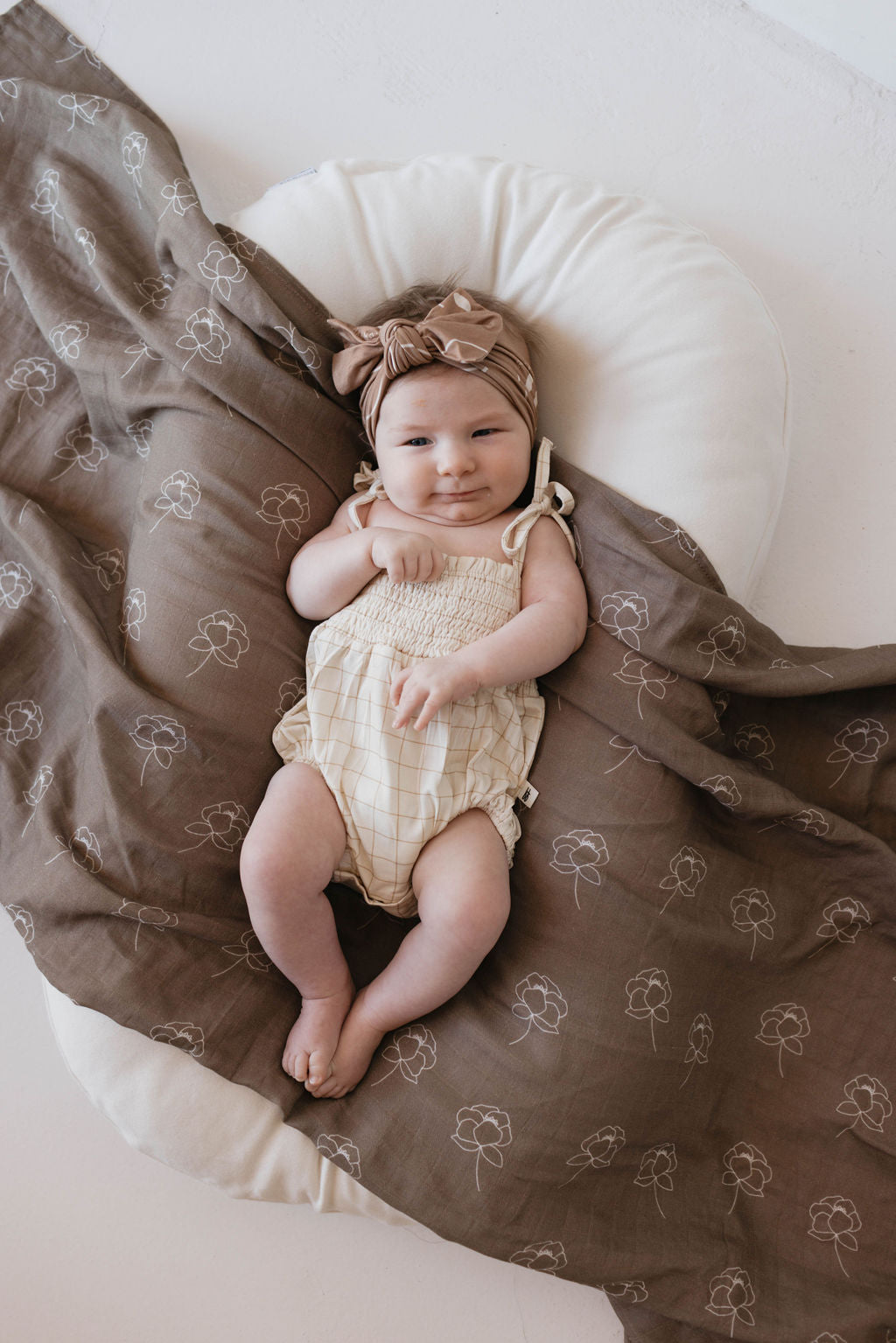 A baby wearing a forever French baby Bamboo Tie Top Romper | Golden Grid with adjustable tie shoulder straps and a matching headband lies on a white cushion. The baby is covered with a brown blanket decorated with floral patterns, looking content with hands near the face and legs slightly bent.