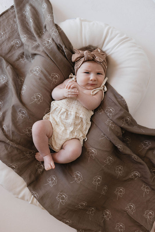 A baby lies on a white cushion, partially wrapped in a brown blanket with simple floral patterns. The baby is wearing a "Golden Grid" Bamboo Tie Top Romper by forever French baby, featuring adjustable tie shoulder straps and paired with a matching beige headband with a bow. The baby has a relaxed expression and is holding their hands together.