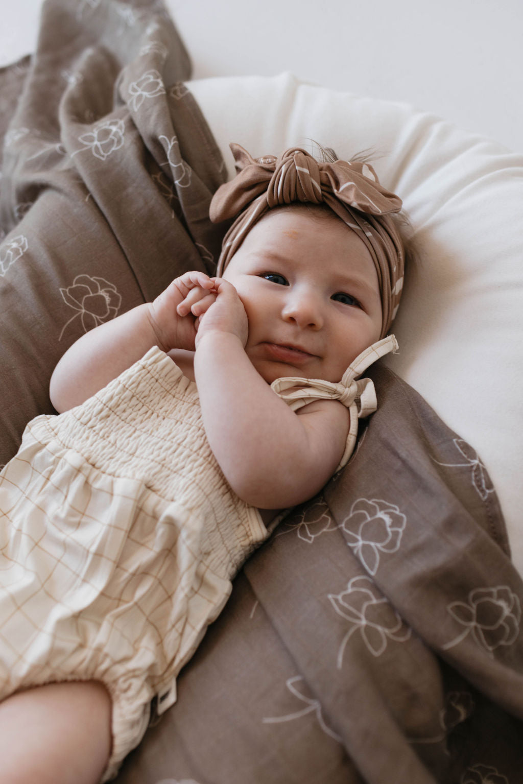 A baby wearing a forever French baby Bamboo Tie Top Romper in the Golden Grid design, complete with adjustable tie shoulder straps and a patterned headband, lies on a soft blanket adorned with floral designs. The contented infant rests on their back with their hands near their face, while the blanket drapes softly around them, creating a cozy setting.