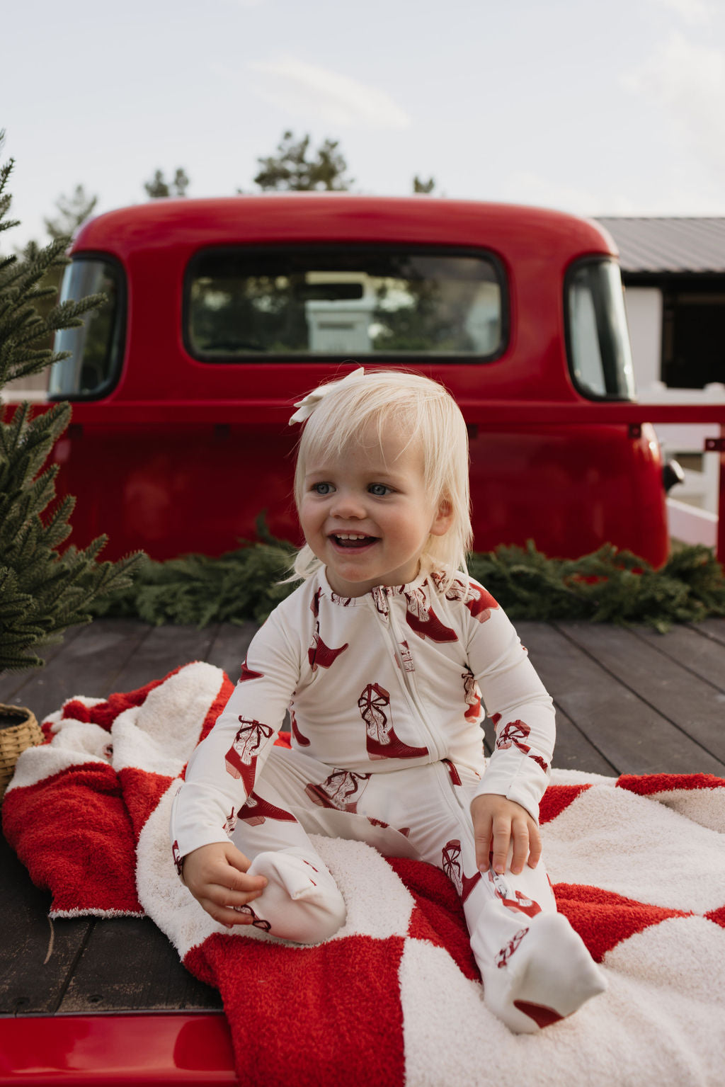 A smiling toddler with blond hair is seated on a red and white blanket in the back of a red truck, wearing lolo webb's festive Bamboo Zip Pajamas in the Cowgirl Christmas pattern. The breathable sleepwear is perfect for play, surrounded by lush greenery.