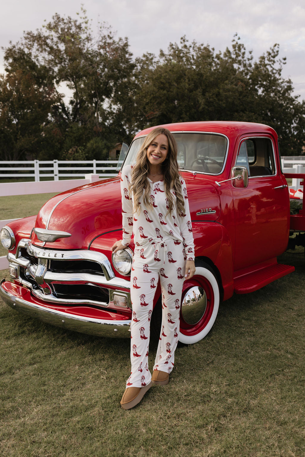 A woman wearing lolo webb's Cowgirl Christmas Women's Bamboo Pajamas, featuring red patterns, stands smiling in front of a vintage red Chevrolet truck on a grassy area, with trees and a white fence in the background.