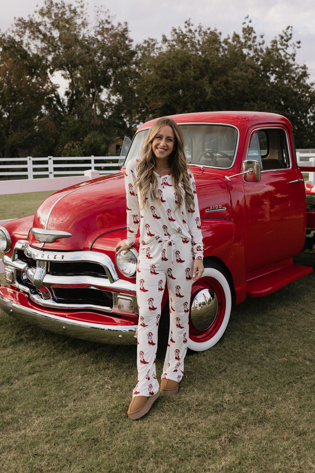 A woman grins while standing in front of a classic red Chevrolet truck, wearing the festive "Women's Bamboo Pajamas - Cowgirl Christmas" by lolo webb, adorned with red patterns. She stands on grass, framed by trees and a white fence, embodying comfort and style.