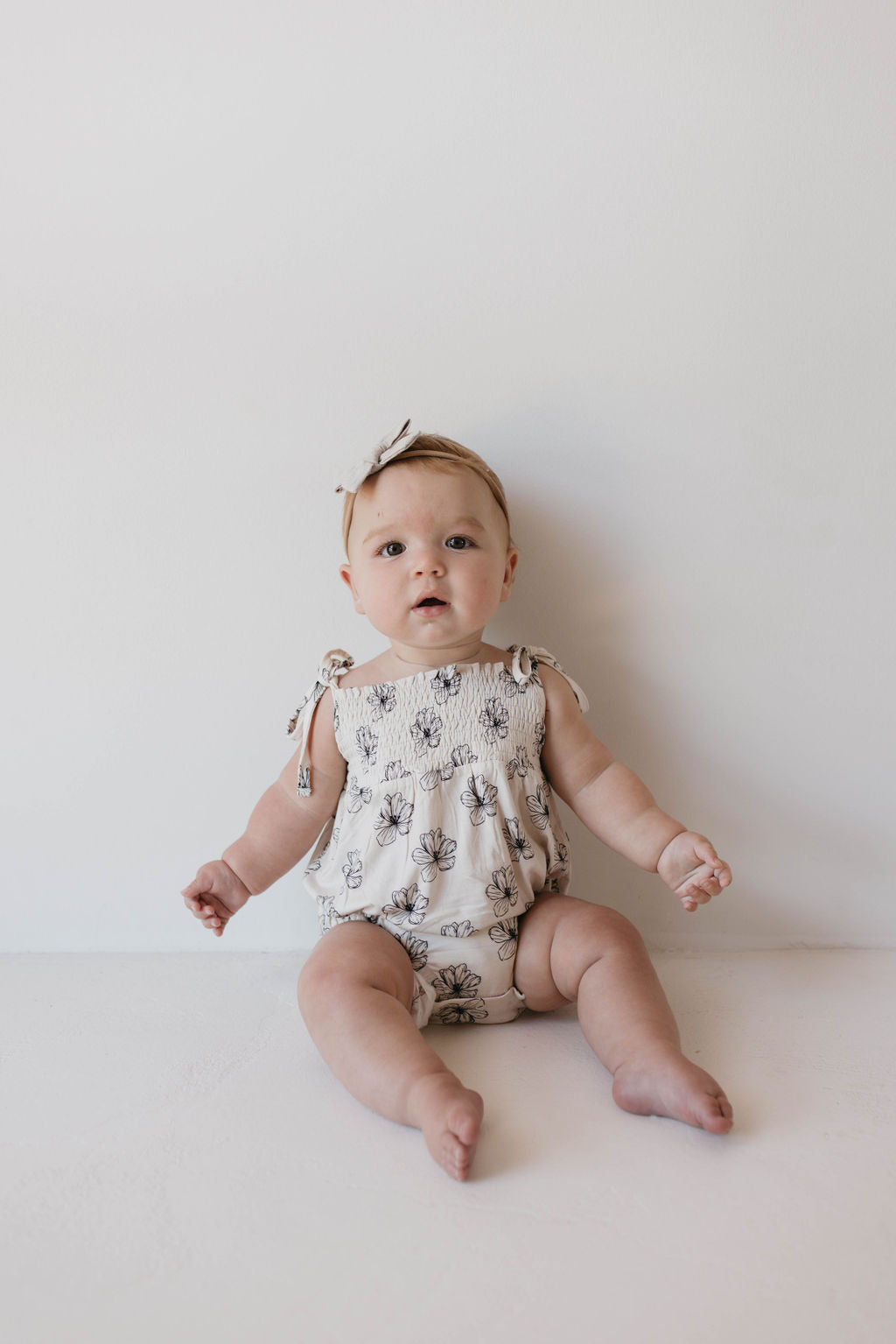 A baby wearing the Bamboo Tie Top Romper in Desert Bloom, a light-colored, floral-patterned outfit with adjustable tie shoulder straps from forever French baby, sits on the floor against a plain white wall. The baby is looking forward with a neutral expression and has a matching bow headband.