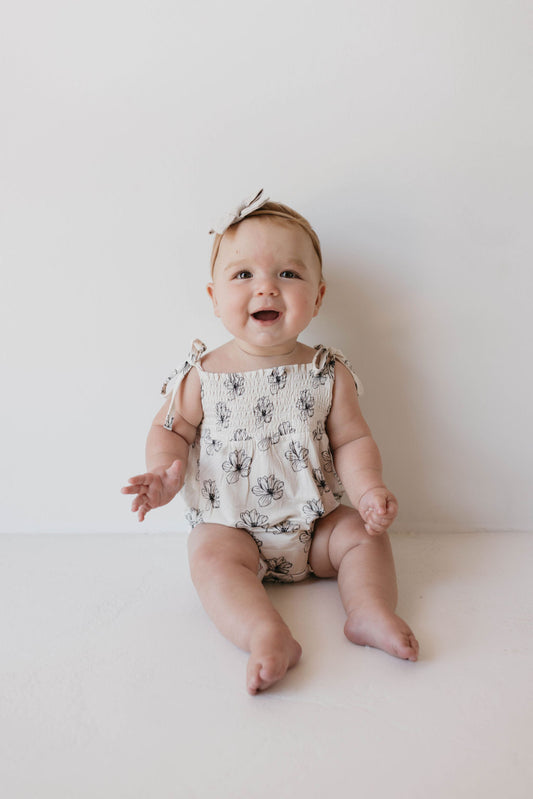 A baby sits on a white surface against a white background, smiling and wearing the *Bamboo Tie Top Romper | Desert Bloom* by *forever French baby*. The romper features adjustable tie shoulder straps. The baby's outfit is complemented with a light-colored bow headband.