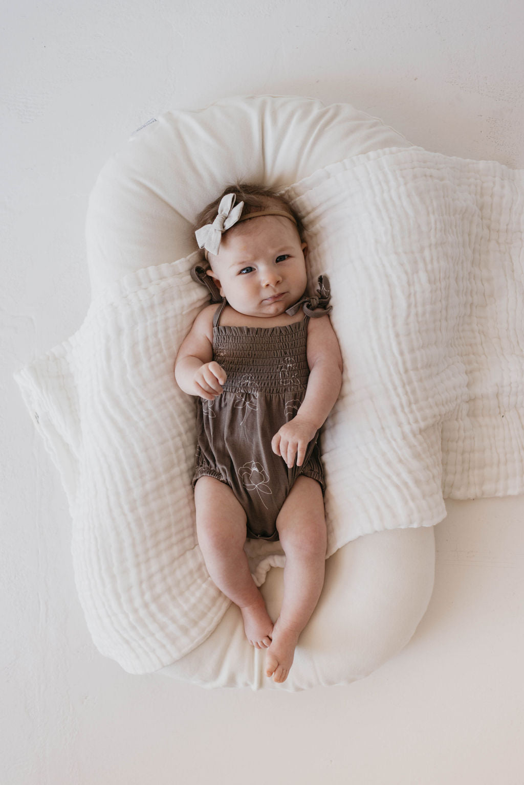 A baby in a Bamboo Tie Top Romper from forever French baby, featuring the Sweet Dreams Floral design with adjustable tie shoulder straps, and a white bow on their head lies on a white, quilted cushion with a matching blanket. The baby has wide, curious eyes and their hands are slightly clenched. The background is a plain, off-white surface.