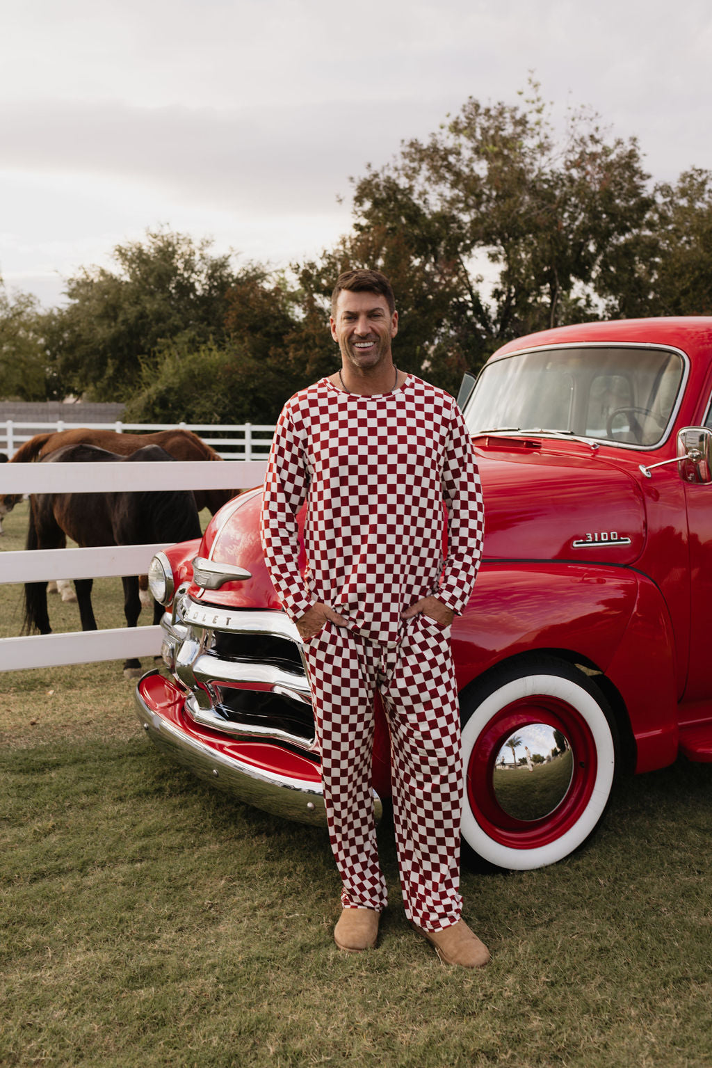 A person stands smiling in front of a vintage red truck, wearing the Quinn bamboo pajamas by lolo webb, featuring a breathable red and white checkered pattern. The outdoor setting includes a white fence and trees in the background, with a horse visible behind the fence.