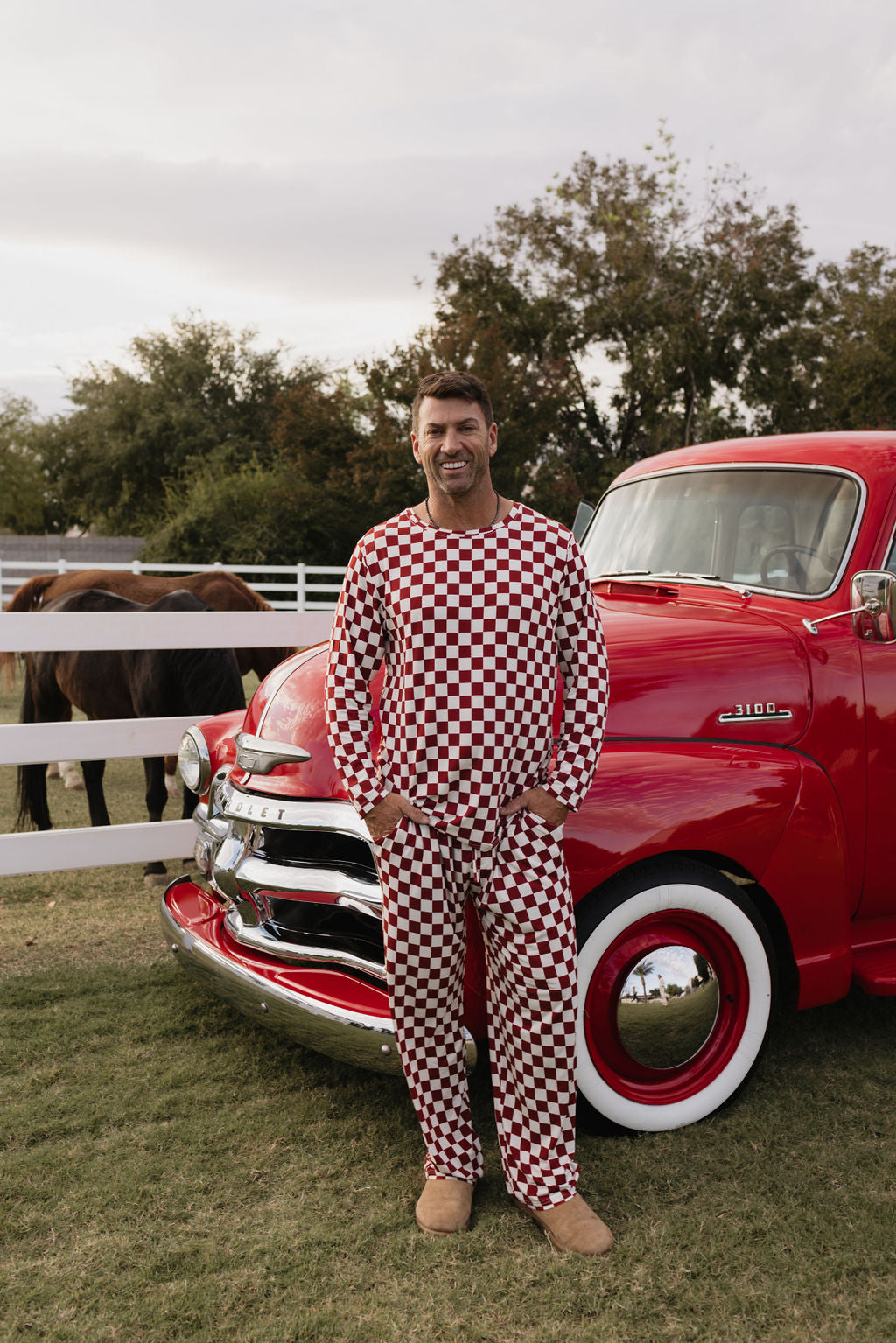 Wearing lolo webb's Quinn men's bamboo pajamas in a striking red and white checkered pattern, a person stands smiling in front of a vintage red truck. A white fence and a couple of horses provide the backdrop on the grassy field, underscoring the breathable fabric that enhances such peaceful moments.