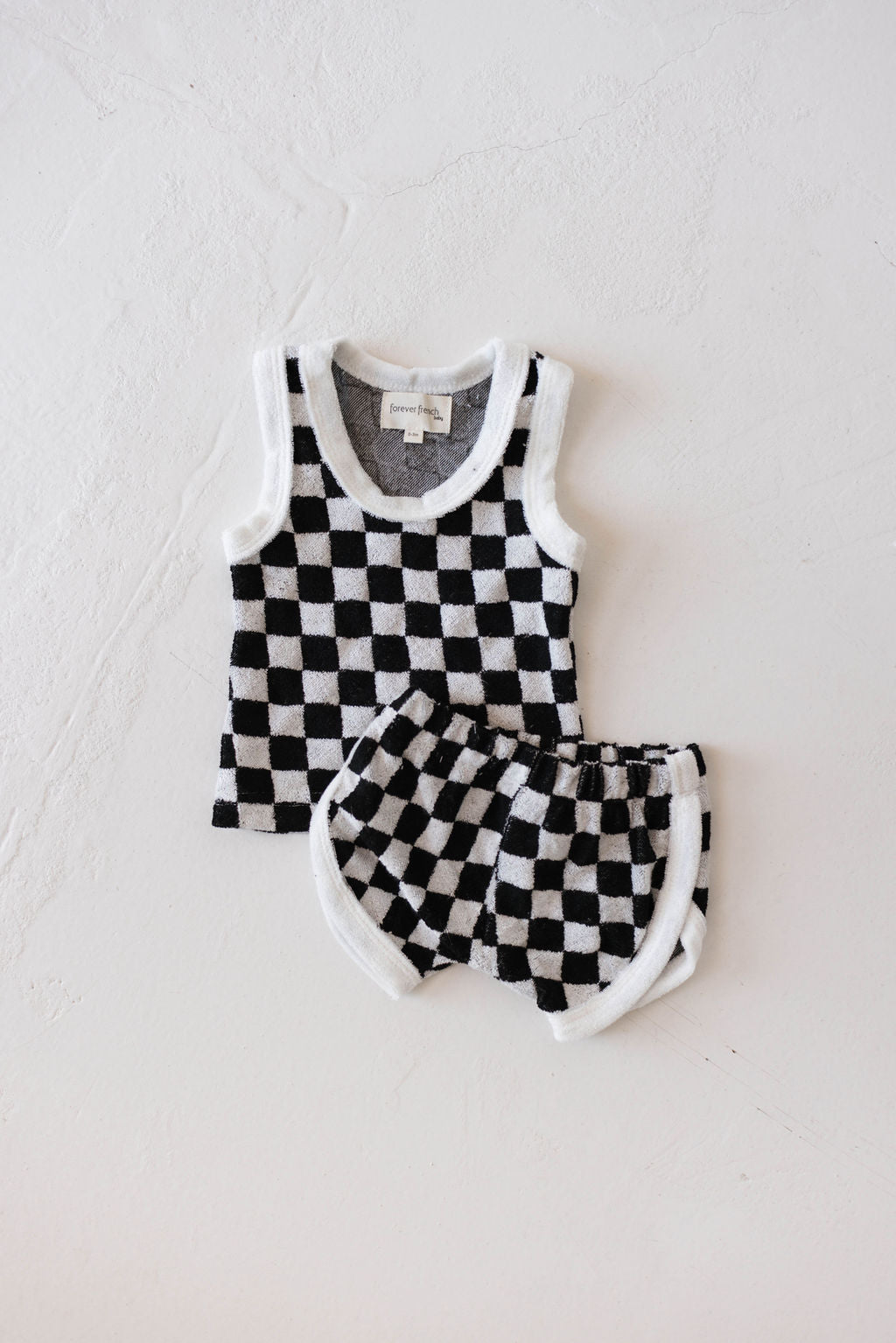 A stylish ensemble highlighting the Forever French Terry Tank Short Set in Black and White Checker, adorned with white trim along the edges. This outfit, featuring a soft woven texture, lies gracefully on a white surface.