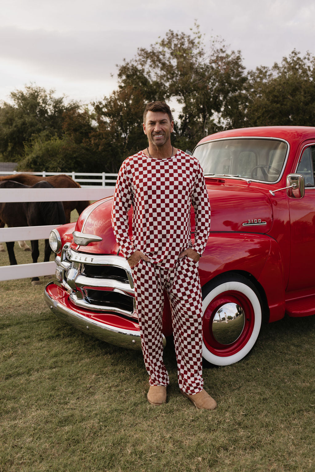 A person wearing the Quinn men's bamboo pajamas by lolo webb, featuring a red and white checkered pattern, stands smiling in front of a vintage red truck adorned with chrome details and whitewall tires. In the background are horses and a white fence on a grassy field, adding to the serene scene.