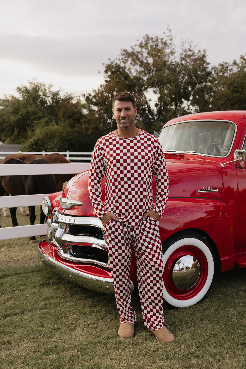 A man in a breathable outfit with red and white checkered patterns stands by a vintage red truck, surrounded by trees and horses, showcasing comfort reminiscent of the Quinn men's bamboo pajamas from lolo webb.