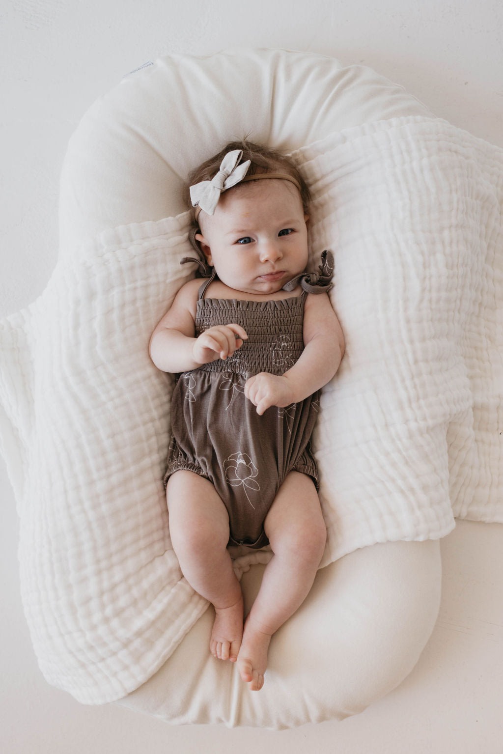 A baby wearing a forever French baby Bamboo Tie Top Romper in the Sweet Dreams Floral pattern, along with a white headband, rests on a cushioned surface. The baby is lying on a soft, neutral fabric background and looking directly at the camera with a calm expression.